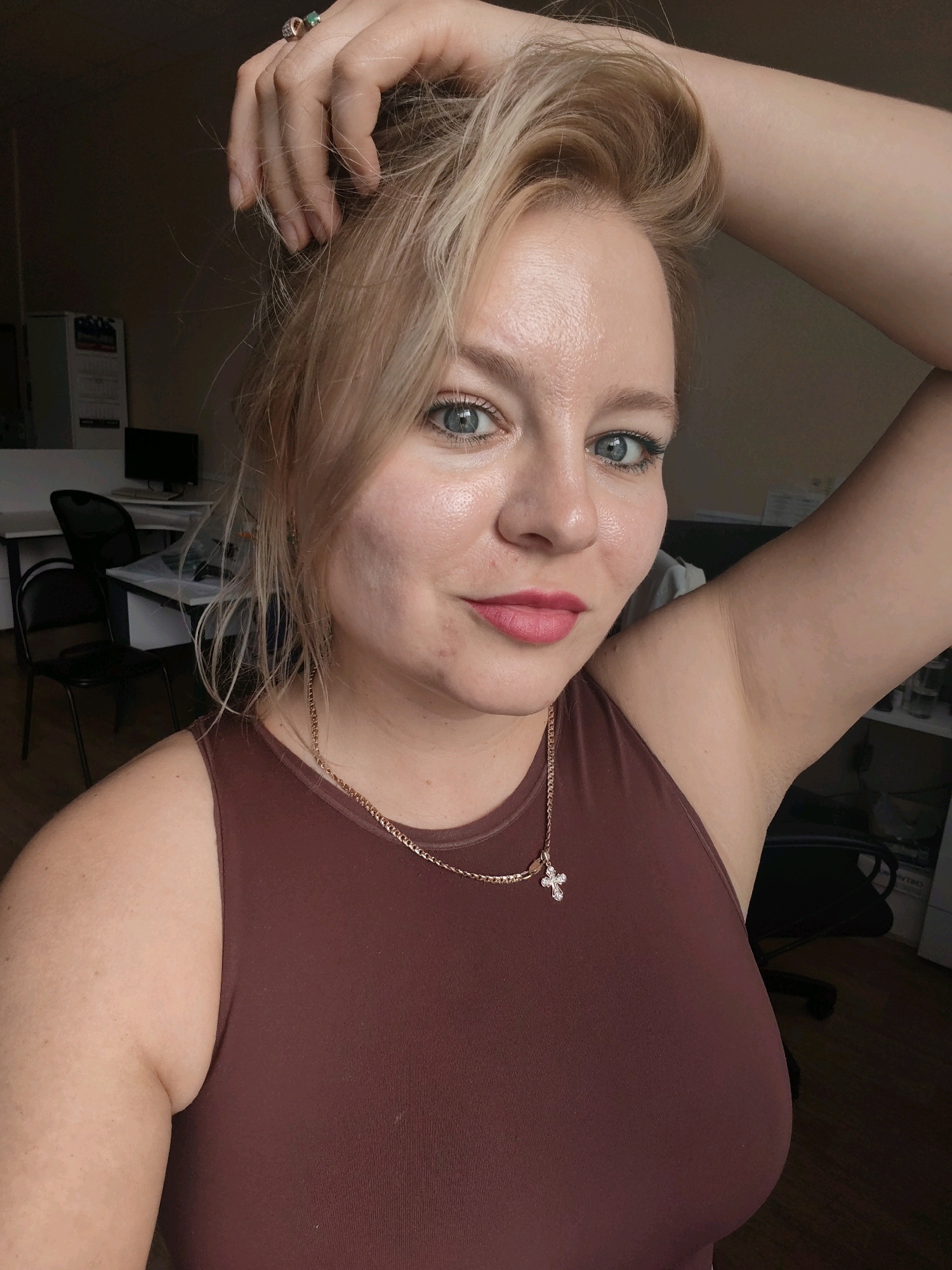 A young woman with blonde hair and blue eyes is taking a selfie. She is wearing a brown tank top and a gold chain with a cross pendant. Her left hand is raised, touching her hair. The background appears to be an office or classroom setting with desks, chairs, and filing cabinets. The lighting suggests an indoor environment, possibly during daylight hours.