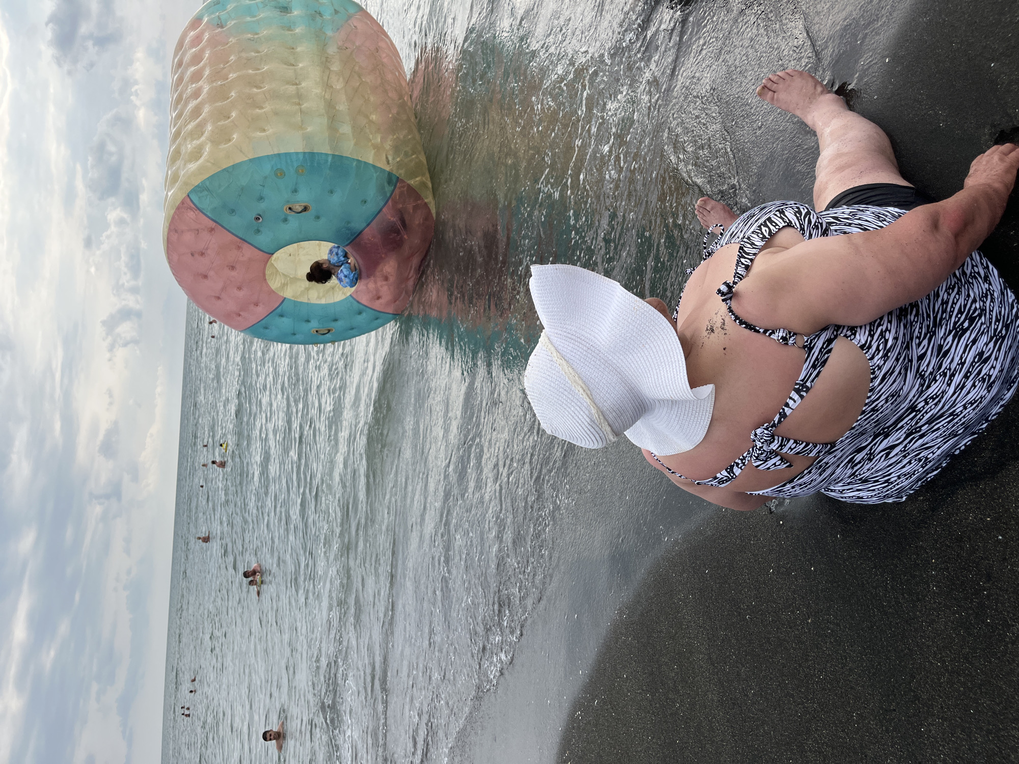 In Ureki, Georgia, at the Black Sea coast, a woman sits on the wet sand near the water's edge, facing away from the camera. She is wearing a white wide-brimmed hat and a black and white patterned swimsuit. Her legs are extended towards the water. In the shallow water, a large, translucent inflatable sphere with sections of pink, blue, and yellow is floating. Inside the sphere, a child wearing a blue life vest is visible. Several other people are swimming and playing in the sea in the background. The sky is overcast with clouds.
