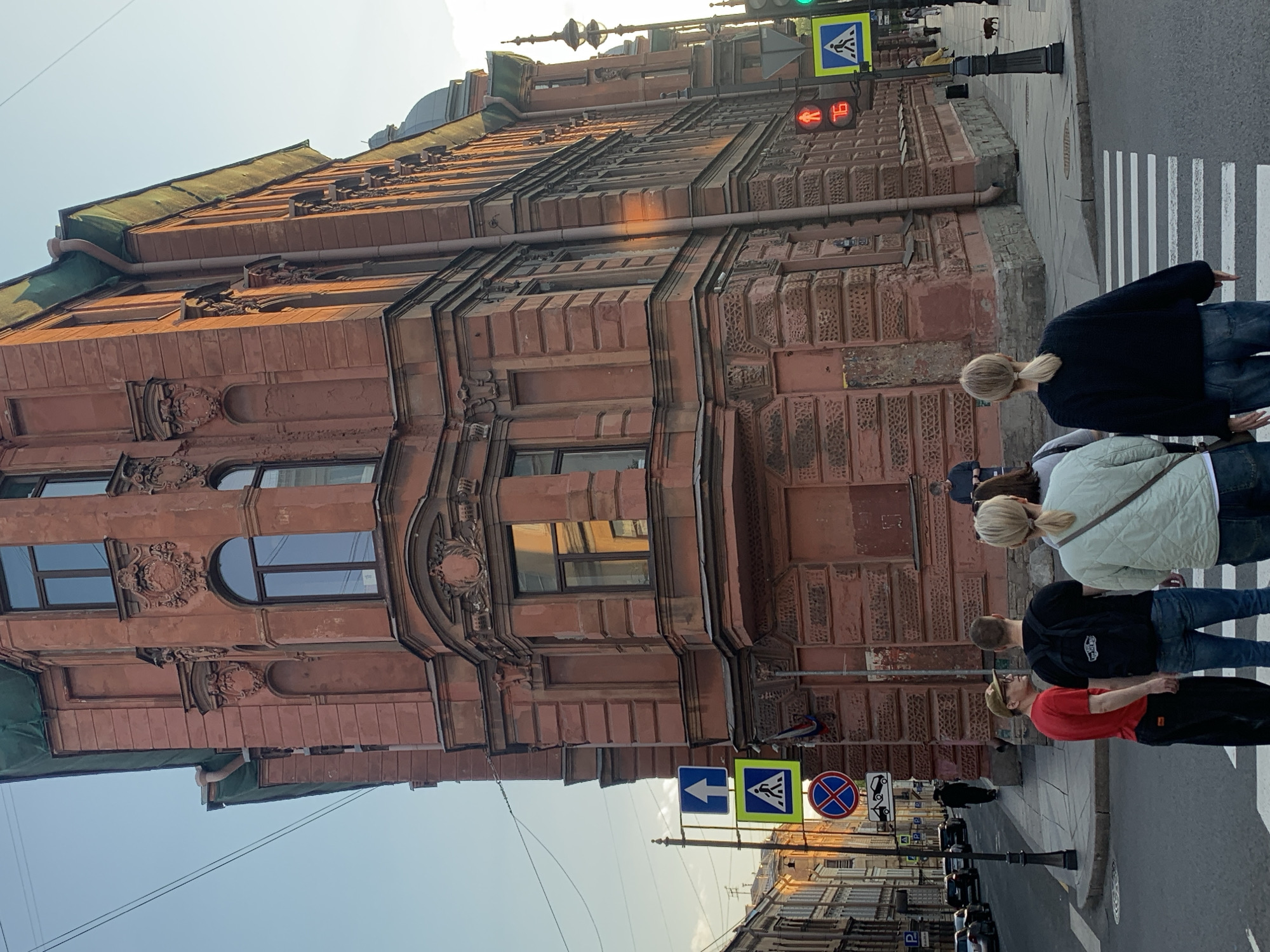 Here is a detailed description of the image:
**Scene**: An outdoor scene in the city of Saint Petersburg, Russia, possibly in a central or historical district. A group of people is crossing a pedestrian crosswalk. The background features classic architecture, with intricate details and a terracotta color scheme.
**Architecture**: The prominent building has multiple floors with ornate window frames, moldings, and detailed facades. The building's architectural style suggests a late 19th or early 20th-century construction period. Some parts of the building have different materials, such as a sheet metal roof.
**People**: The group of people in the foreground is walking across the crosswalk. Their hairstyles are visible, and their clothes suggest casual attire.
**Objects**: Street signs, including pedestrian crossing signals, are visible near the crosswalk. A pedestrian signal shows a red light. Street lamps are positioned at the edge of the sidewalk. There are vehicles parked along the street, and some are in motion.
**Environment**: The sky is visible, suggesting daylight. The lighting conditions indicate it might be late afternoon or early evening.