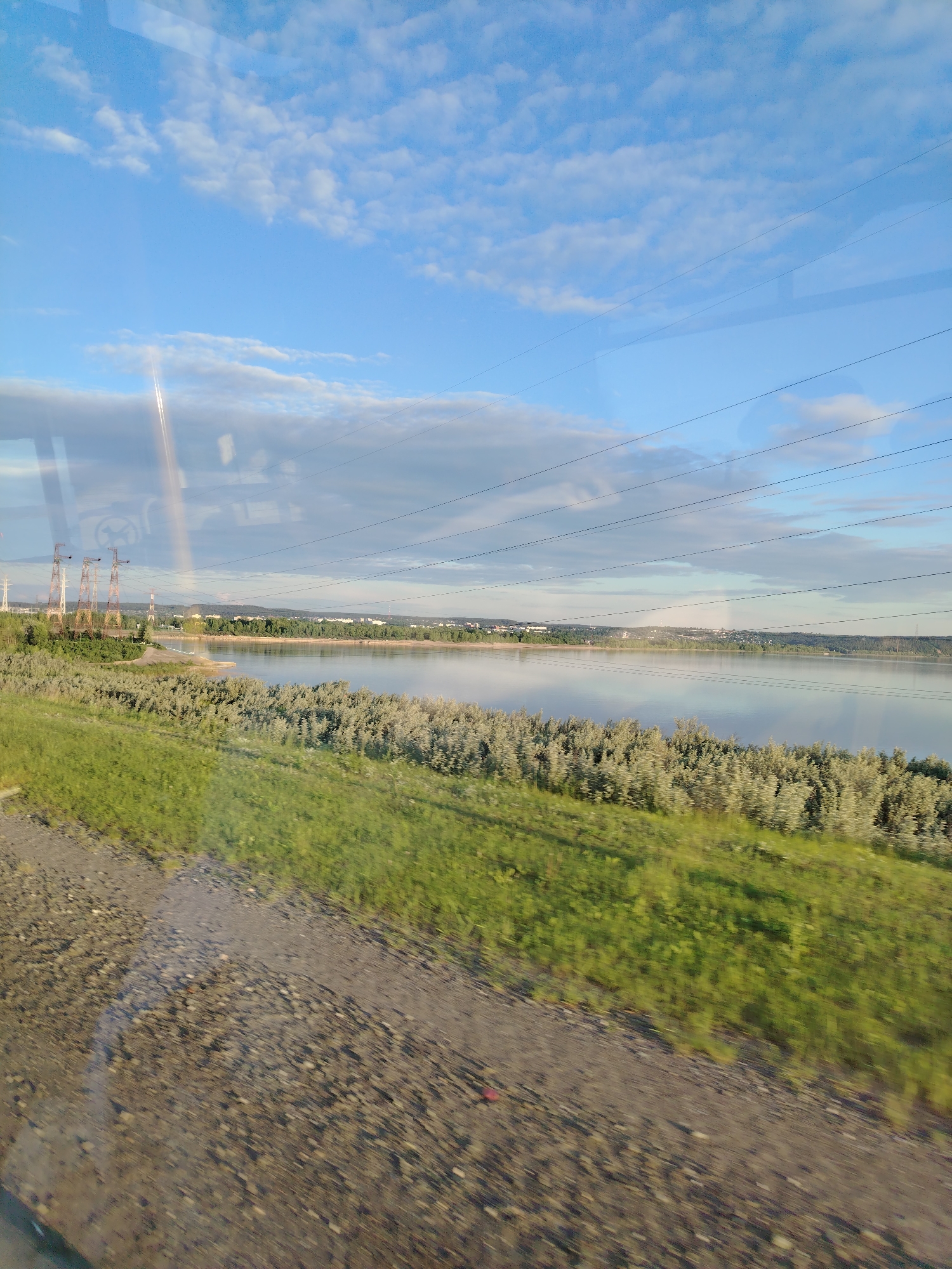 The image depicts an expansive outdoor landscape, likely situated in the city of Chaikovsky, Russia, viewed through a window, evidenced by visible reflections. The upper portion of the frame features a bright blue sky with scattered white cumulus clouds, along with some lower, more diffused greyish clouds. Multiple thin, dark horizontal lines, identified as power lines, traverse the sky and are also visible as reflections across the upper half of the image. A prominent, bright vertical streak, indicative of a strong light reflection, bisects the left side of the sky and extends into the water.
The middle ground is dominated by a wide, placid body of water, possibly a river or reservoir, reflecting the sky and clouds. Along the distant shoreline of this water body, a dense band of light green, deciduous bushes or small trees is visible. Beyond these, a darker band of forested land leads to a distant horizon where the silhouette of a city or town with various buildings is discernible. On the left side of the distant land, several large, rust-colored electricity pylons or transmission towers with intricate lattice structures are present.
The foreground of the image consists of a blurry, verdant green grassy area on the right, transitioning to a gravel or dirt path/road on the left, with some reddish debris visible on the path. The blur in the foreground suggests the image was captured from a moving vantage point, such as a vehicle. No individuals or specific actions are identifiable within the scene.