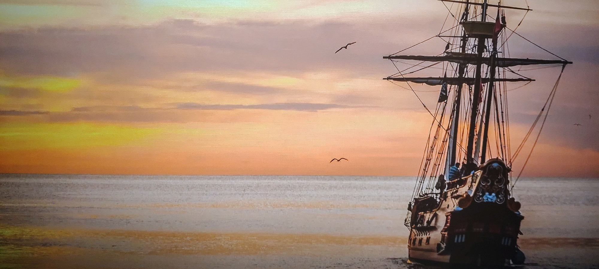This image depicts a tall ship sailing on the ocean at sunset. The sky is filled with warm, soft colors ranging from yellow and orange to pink and purple, with scattered clouds. The ocean surface is calm, reflecting the colors of the sky. The ship is viewed from the stern, showing its intricate wooden details and rigging. Two seagulls are visible in flight against the sunset sky. The scene evokes a sense of peace and adventure. There is no identifiable connection to Hyderabad, India, in this image.