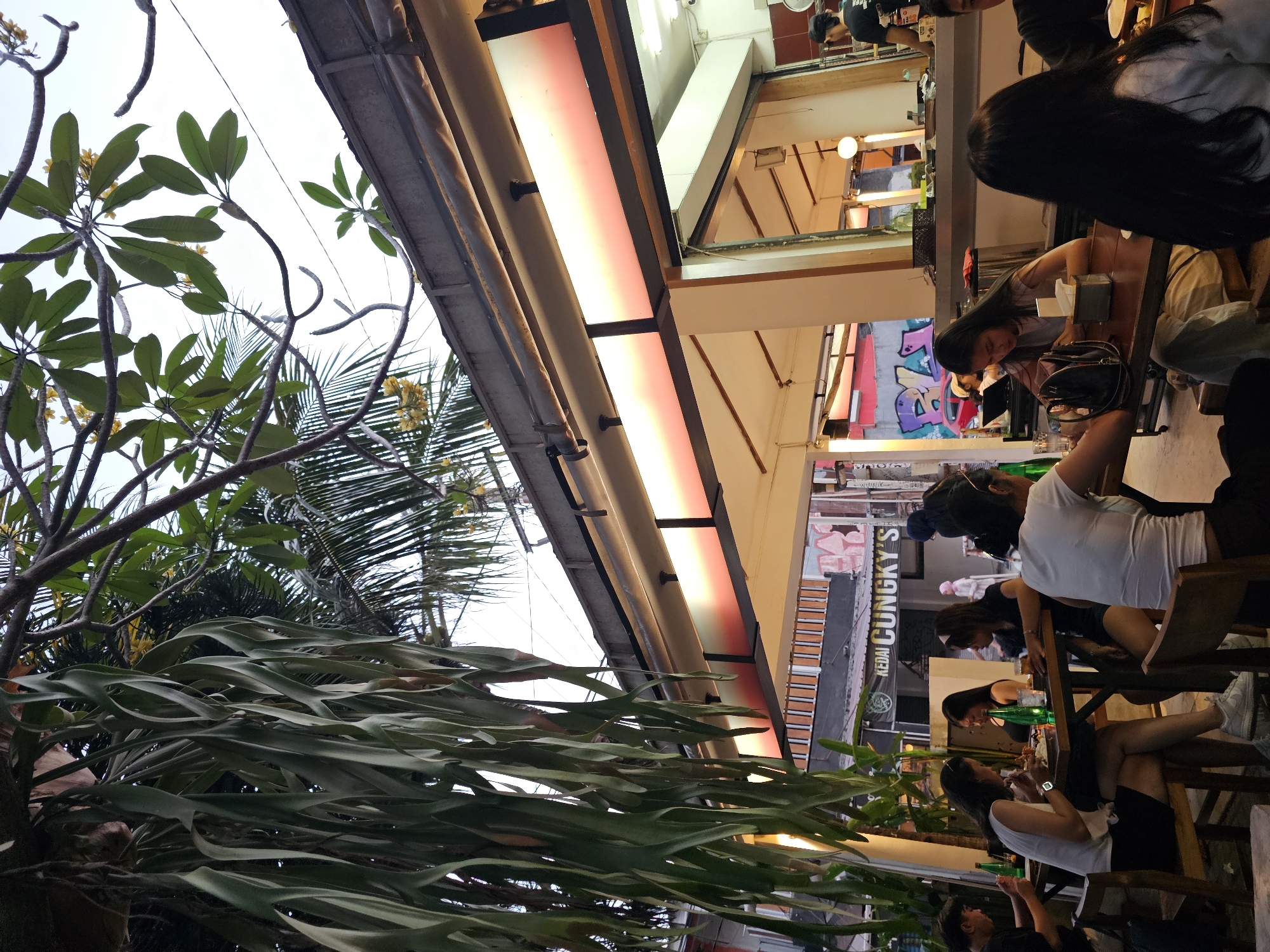 The image shows an outdoor seating area of a restaurant in Yogyakarta, Indonesia. The restaurant appears to be a casual eatery with wooden tables and chairs. Several people are seated at the tables, eating and socializing. There are plants hanging from above, creating a natural and lush atmosphere. The background shows buildings and street scenes, indicating an urban environment. The overall impression is that of a lively and pleasant dining experience.