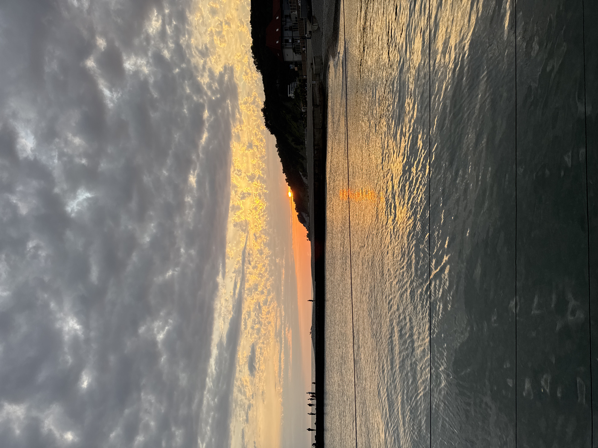 The image depicts a serene sunset scene over the Black Sea in Sochi, Russia. In the foreground, the calm water reflects the warm hues of the setting sun and the cloudy sky. The sky is filled with a mix of dark, moody clouds and patches of vibrant orange and yellow light where the sun is setting. A man-made pier or seawall stretches horizontally across the middle ground, silhouetted against the bright sky. Several small figures of people, appearing as dark silhouettes, are standing on this structure, some appearing to be fishing. On the right side of the image, a treeline obscures a hillside, upon which several buildings with red roofs are visible. The overall impression is one of a peaceful evening at the coast.