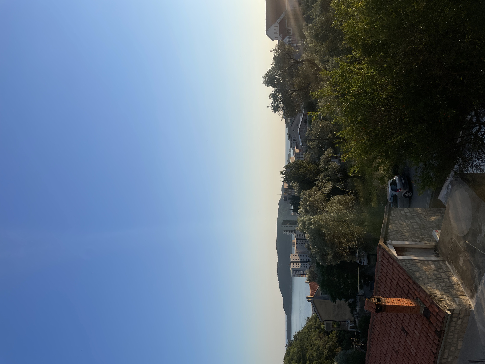 The image depicts a view of the city of Rafailovići, Montenegro, as seen from an elevated vantage point. The sky is a clear, vibrant blue, dominating the upper portion of the frame. Below, a body of water, likely the Adriatic Sea, stretches towards a distant, hazy coastline. In the middle ground, a cluster of buildings, including a multi-story apartment complex, are visible amidst lush green foliage and trees, many of which appear to be olive trees. The architecture suggests a Mediterranean setting, with tiled roofs and stone accents on some of the structures. A small, silver car is parked on a road in the middle distance. The foreground features the roof of a building with red tiles and a brick chimney, as well as a stone wall with a wooden door. Sunlight casts dappled shadows on the scene, indicating it was taken during the late afternoon or early morning. There are no people or animals visible in the image.