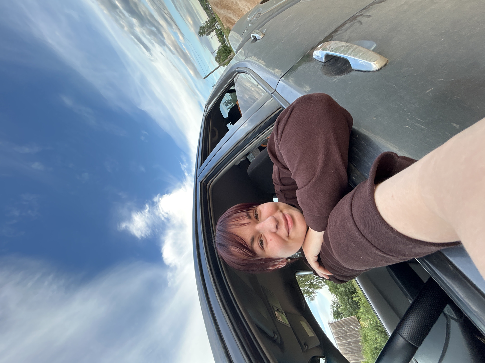 The image shows a person leaning out of a car window. The person has short, dark hair and is wearing a brown hooded sweatshirt. They are smiling at the camera, and their arm is resting on the car door. The car appears to be parked outdoors, with a cloudy blue sky in the background. There are trees and a building visible in the distance. The time of day appears to be daytime, and the weather seems to be partly cloudy. The image is a selfie, taken from a slightly low angle.