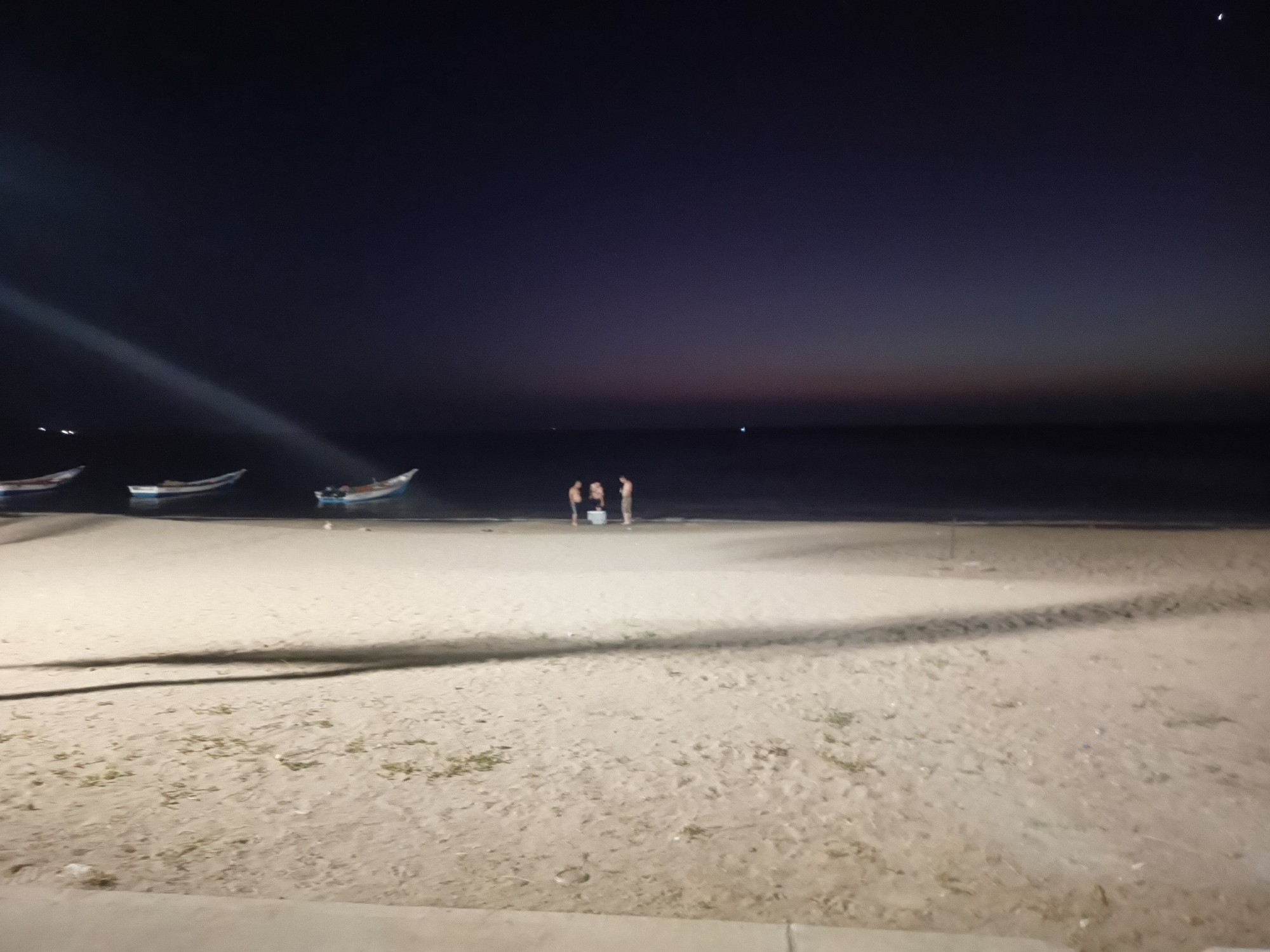 The image depicts a nighttime beach scene in Juan Griego, Venezuela. In the background, the sky transitions from a dark blue to orange at the horizon. The sea is visible. Three fishing boats are anchored near the shore on the left. A group of three people stand near the water's edge. The foreground shows a sandy beach. Shadowy shapes cross the foreground, possibly from vegetation or structures out of frame.