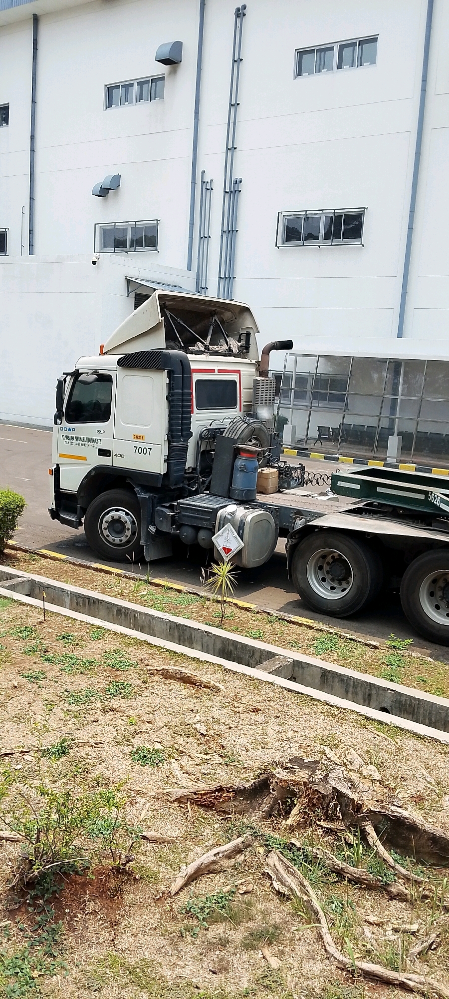 An outdoor daytime, medium-shot, vertical-orientation photograph captures a white tractor-trailer truck parked on asphalt in what appears to be an industrial area. The truck, identified by the number 7007 and the logo "DOWA," is positioned at an angle facing left, with its cab area slightly elevated. The truck's front tire is turned left, and the vehicle is empty, suggesting it is either awaiting a load or has recently completed a delivery. The truck is situated in front of a large white building with numerous windows and grey ductwork visible on its facade. The foreground of the image shows a dry, grassy area with a concrete drainage ditch. The overall scene conveys a sense of industrial activity or storage. The building's architecture and the dry climate suggest the location could be Depok, Indonesia, a city known for its industrial zones and tropical weather.