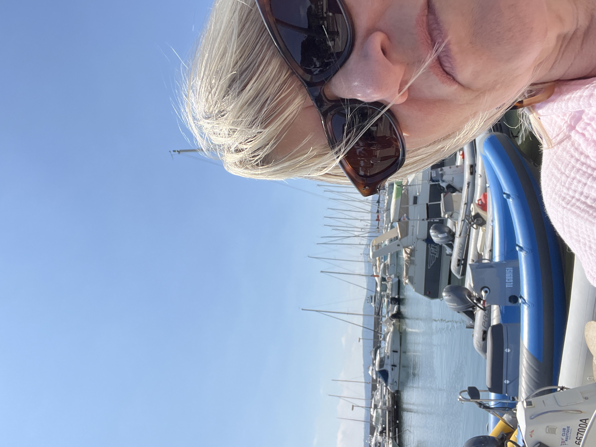 The image captures a woman with blonde hair and sunglasses, possibly in Sainte-Maxime, France. She's likely on a boat or near a marina, as several boats and masts are visible in the background. The sky is clear and blue, suggesting a sunny day. In the foreground, part of a blue and gray inflatable boat is visible, and other boats are docked nearby. The image is taken from a close, slightly angled perspective focusing on the woman's face.