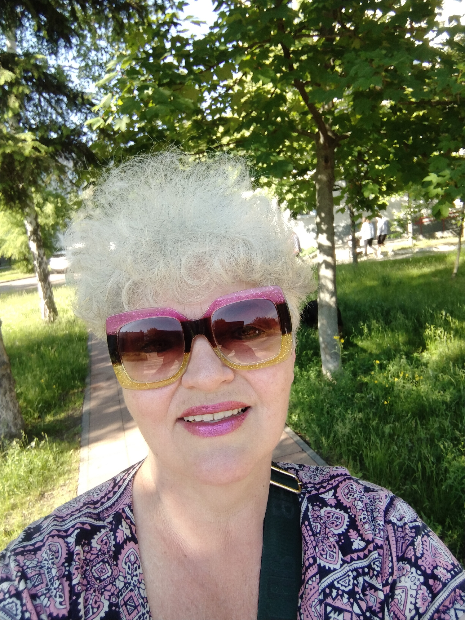 A close-up selfie captures a smiling woman outdoors in what appears to be a park or green area in Horishni Plavni, Ukraine. The subject, an older woman, features voluminous, curly white hair. She is wearing large, square-framed sunglasses with a distinctive glittery design, transitioning from pink on the top to gold on the bottom. Her lips are adorned with bright, glossy pink lipstick, and she has a pleasant, gentle smile. She is dressed in a patterned top with intricate designs in shades of pink, purple, white, and black, and a dark green strap, possibly from a bag, is visible on her right shoulder.
The background depicts a sunny outdoor setting with lush green foliage. Trees, including both deciduous types with broad leaves and some coniferous trees, are visible. Bright sunlight filters through the leaves, creating dappled light and shadows on the ground. A paved pathway is seen to her left and receding into the background. Areas of green grass, some with small yellow flowers, line the path. In the deeper background, blurry figures of other people can be observed walking, suggesting a public recreational space. The bright lighting indicates it is daytime with clear, sunny weather. There is no visible text in the image.