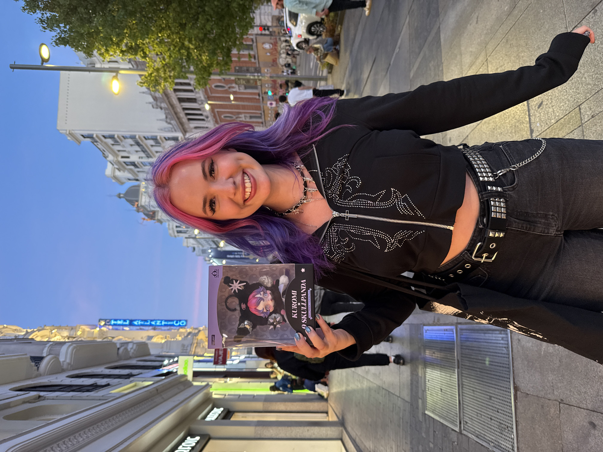 A young woman with pink and purple hair is standing on a city sidewalk, holding a "Kuromi x Skullpanda" figurine in its original packaging. She is smiling and holding up the figurine towards the camera. She is wearing a black cropped jacket with studded detailing, black jeans with a studded belt, and a silver necklace. The background shows a city street at dusk or early evening. The buildings have ornate architecture, and there are streetlights and a neon sign visible. People can be seen walking in the background. The sign reads "Hotel Atlantico," and the architecture identifies the location as the Gran Vía in Madrid, Spain.