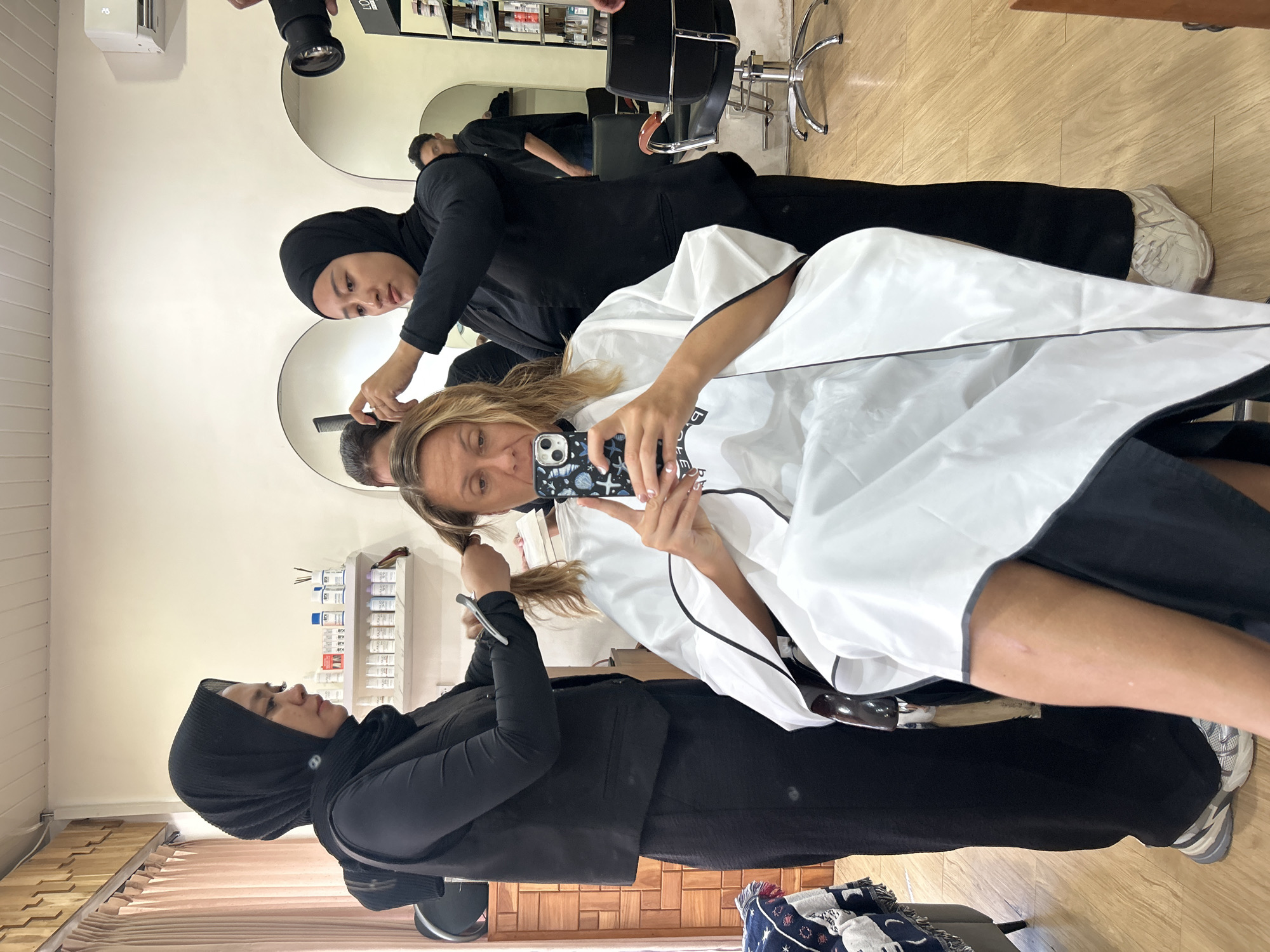 In North Kuta, Indonesia, a Caucasian woman is seated in a salon chair, draped in a white salon cape, and taking a selfie with her phone. Her blonde hair is being styled by two women wearing black hijabs and black salon attire. The woman on the left is holding scissors and a comb, while the woman on the right is holding a comb to the client's hair. In the background, a man in a black shirt and black pants stands near a mirror, and shelves stocked with hair products are visible. The salon appears to be well-lit with natural light coming from the left side of the image, and the floor is made of light-colored wood.