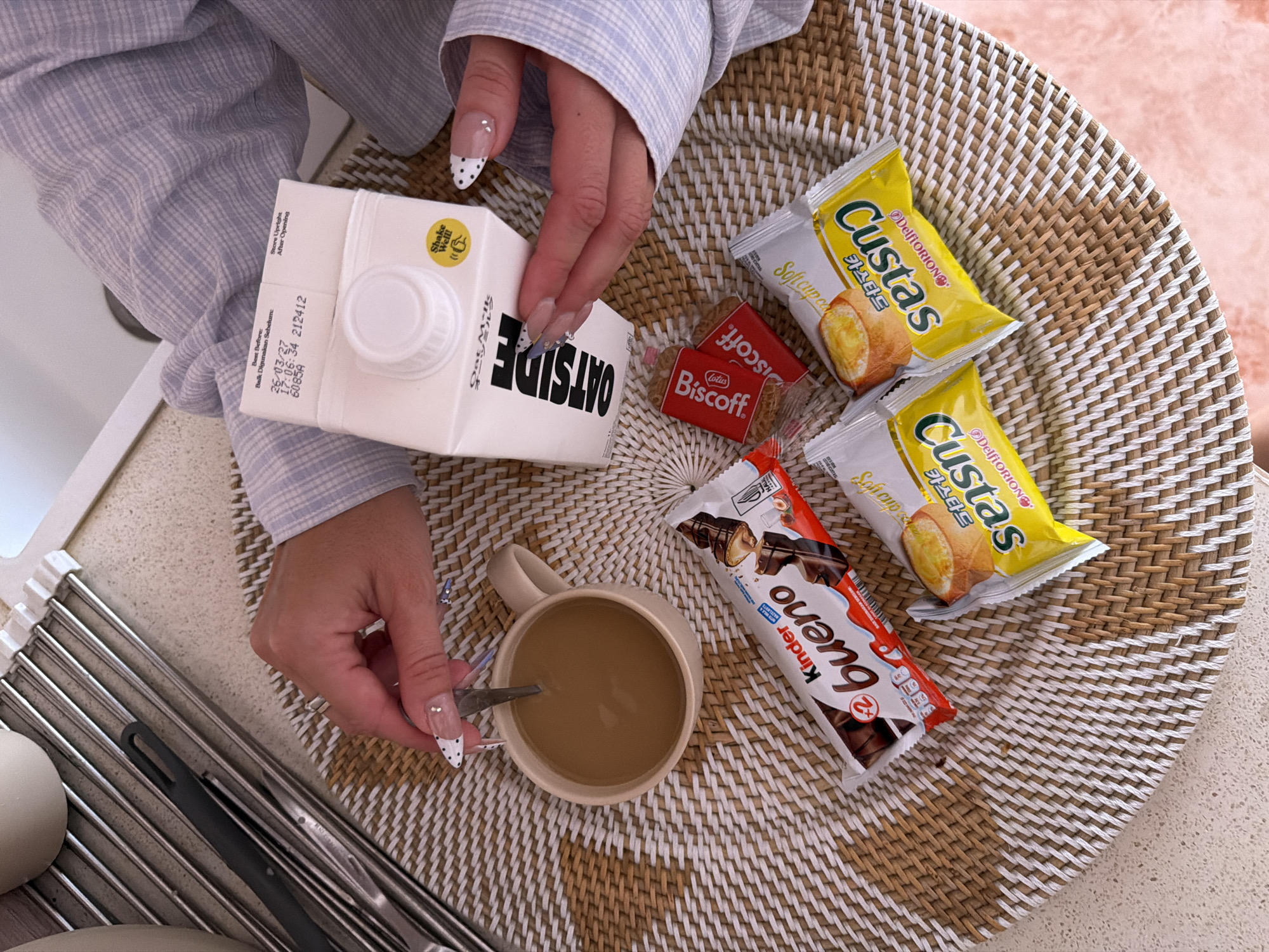 The image depicts an aerial view of a breakfast arrangement. A woman, wearing a striped shirt, is stirring a cup of coffee. Beside the cup are two individually wrapped Custas cakes, a Lotus Biscoff biscuit, and a Kinder Bueno chocolate bar. Adjacent to these items is a carton of Oatside oat milk. A dish rack containing utensils is partially visible at the bottom left corner of the image. The breakfast is arranged on a woven circular mat. The image was taken in North Kuta, Indonesia. The carton of oat milk has a label that indicates the date and time it was processed.