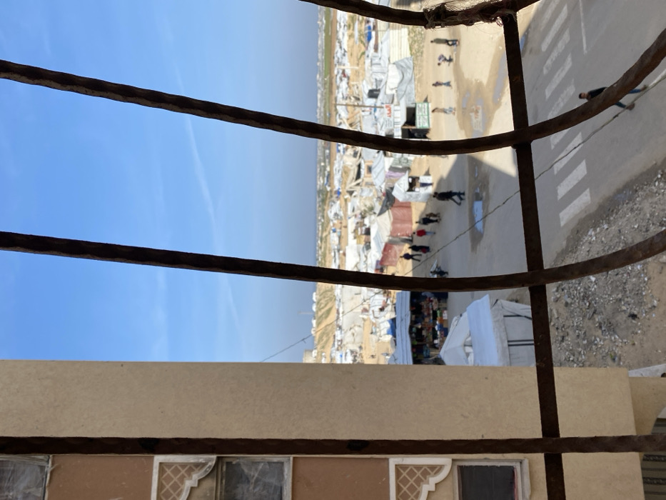 The image, taken from an elevated perspective through a metal grate, depicts a densely populated temporary settlement in Khan Yunis, Palestinian Territory. In the left foreground, a section of a beige building with two small, damaged windows and a decorative relief panel is visible. Dark, rusty vertical and horizontal metal bars, likely from a window grate or balcony railing, frame the scene.
Below the bars, an asphalt road with a white-striped crosswalk is visible, featuring several muddy puddles. Approximately fifteen individuals, including adults and children, are walking on the road and congregating near makeshift structures. On the left side of the road, a market stall, constructed with tarpaulins and metal poles, displays an assortment of goods, possibly produce. Another structure nearby has Arabic text on its side, reading "الماء" (water).
The midground and background are dominated by an extensive camp composed of numerous temporary shelters, primarily constructed from white, blue, and brown tarpaulins stretched over frames. These shelters are tightly packed and extend far into the distance. Beyond the camp, undulating earthen mounds and sparse, dry vegetation define the horizon under a clear blue sky with faint contrails.
