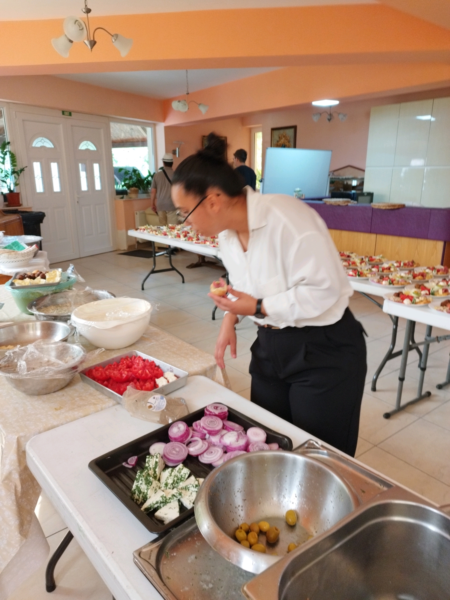 The image depicts a catering event or food preparation area in what appears to be a community hall or restaurant in Bucharest, Romania. In the foreground, a woman wearing a white shirt and black pants is visible from the waist down, holding a small pastry or piece of food in her right hand and examining it. She is standing near a table laden with food items.
On the table, there are several trays and bowls containing prepared food. One tray holds chopped tomatoes and cubed white cheese with herbs. Another black rectangular tray contains sliced red onions and more cubed white cheese with herbs. A metal bowl in the lower right foreground contains green and brown olives. To the left, other bowls are visible, some covered with plastic wrap. In the background, tables are set up with numerous small appetizers, each artfully arranged. Two men are also present in the background. One man in a light-colored shirt and hat is standing near a doorway, while another man in a dark shirt is further in the background.
The environment suggests an indoor event space with light-colored walls and a patterned floor. A doorway with glass panels and a sign that appears to read "EXIT" is visible on the left side. Decorative lights are attached to the ceiling. The overall scene conveys a sense of preparation and presentation of food for an event.