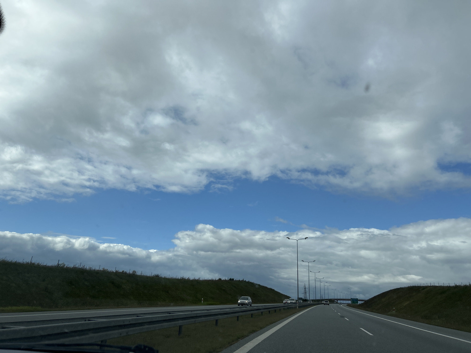 The image depicts a highway scene in Świebodzin, Poland, under a partly cloudy sky. The highway has multiple lanes and is bordered by grassy embankments. Several cars are visible on the road, and streetlights line the side of the highway. The view is from inside a vehicle, suggesting it was taken while driving. The sky is a mix of blue patches and large, grey clouds.