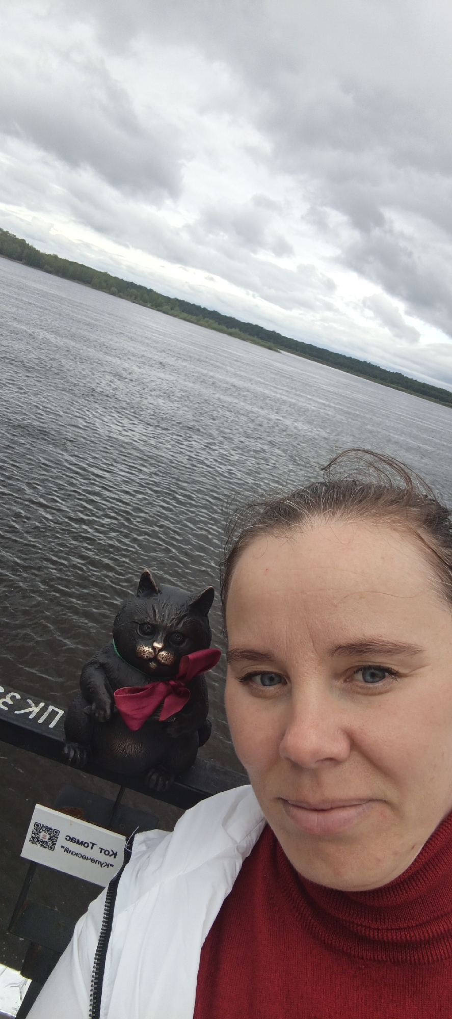 An outdoor close-up selfie captures an adult female and a cat statue positioned on a railing, with a wide body of water and a distant forested shoreline under an overcast sky in the background. The female, occupying the lower right and center of the frame, has light skin, blue eyes, and light brown hair partially pulled back. She wears a white zippered jacket over a red turtleneck. Her gaze is directed towards the camera, maintaining a neutral expression. To her left, a dark, metallic-toned cat statue with a prominent muzzle and an alert expression sits on a dark railing. The statue wears a reddish-maroon bow tie. Below the cat, a white sign featuring a QR code and reversed Russian text is visible, reading "Кот Томас" (Cat Thomas) and "Купчевский" (Kupchevsky). White Cyrillic letters "ЦК 3" are also present on the railing. Beyond the railing, a large body of dark grey water shows ripples across its surface. A distant landmass, covered in dense green trees, forms the horizon. The sky is uniformly covered with thick, grey clouds. This scene is located in the city of Sarapul, Russia.