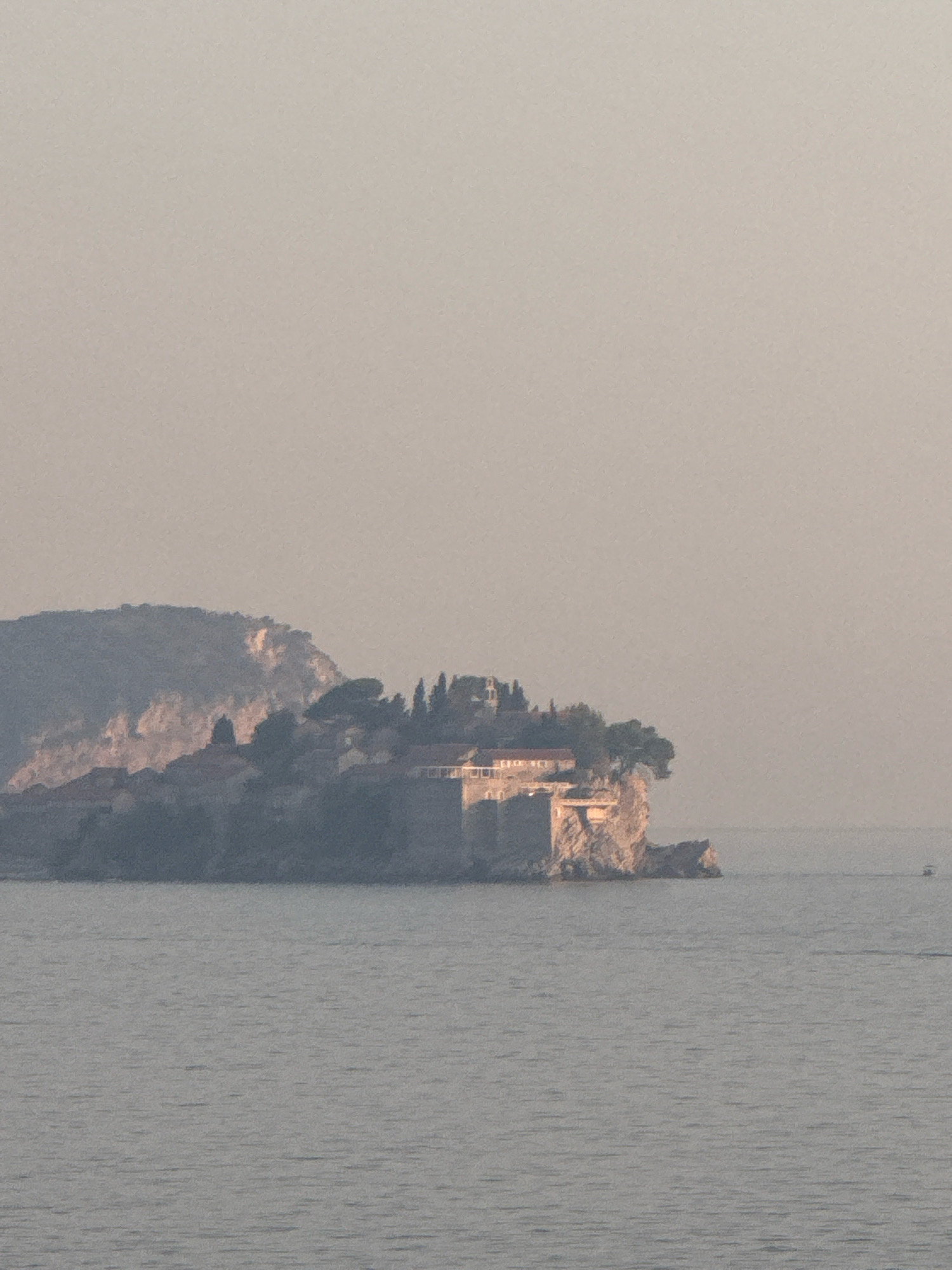 This image shows a view of the coast near Kamenovo, Montenegro, at sunset or sunrise. The foreground is dominated by a calm body of water, reflecting the soft, hazy light of the sky. In the mid-ground, a rocky island juts out of the water, topped with buildings and trees. The island appears to be a historic settlement, with stone structures and a cluster of cypress trees. A small boat is visible in the distance, moving across the water. The background consists of a larger, mountainous landmass, partially obscured by mist or haze. The overall atmosphere is serene and picturesque.