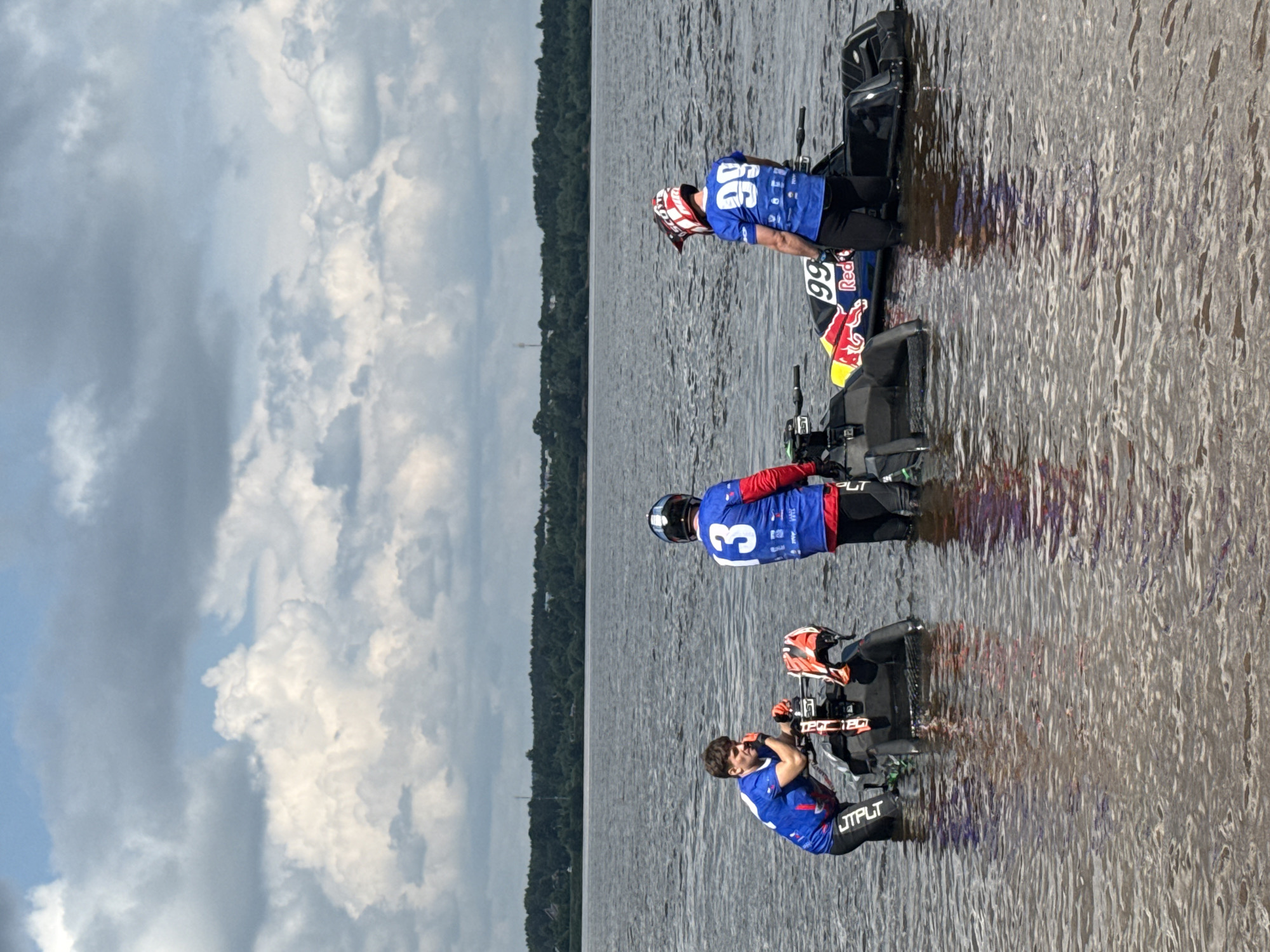 Three individuals, dressed in racing attire, stand in shallow water with jet skis. Two of the individuals are wearing helmets and racing suits with numbers on the back: one with "13" and the other with "99". The jet skis are black with colorful accents, one featuring the "Red Bull" logo. The individuals appear to be preparing for or recovering from an activity, with one looking towards the water and the others looking away from the camera. The environment is a body of water, likely a river, with a treeline and buildings visible in the distance, suggesting an outdoor location near a city. The sky is overcast with dramatic clouds. Based on the context provided, this scene is located at the Kama River in Perm, Russia.