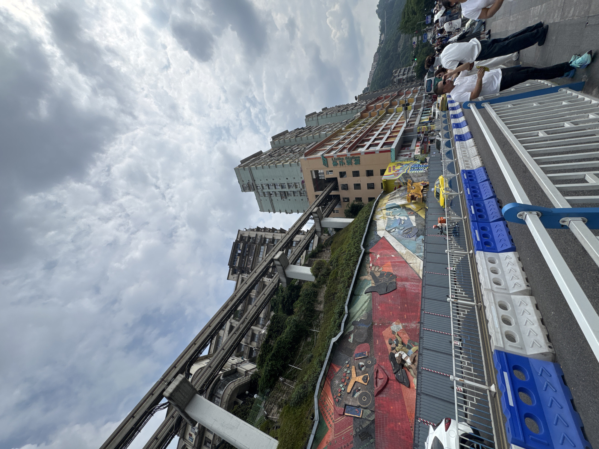 The image depicts a section of the Liziba metro station in Yuzhong District, Chongqing, China. The station runs through a residential building. In the foreground, barriers and people are visible on a platform. The background shows the exterior of the building. A mural adorns a retaining wall. The sky is overcast.