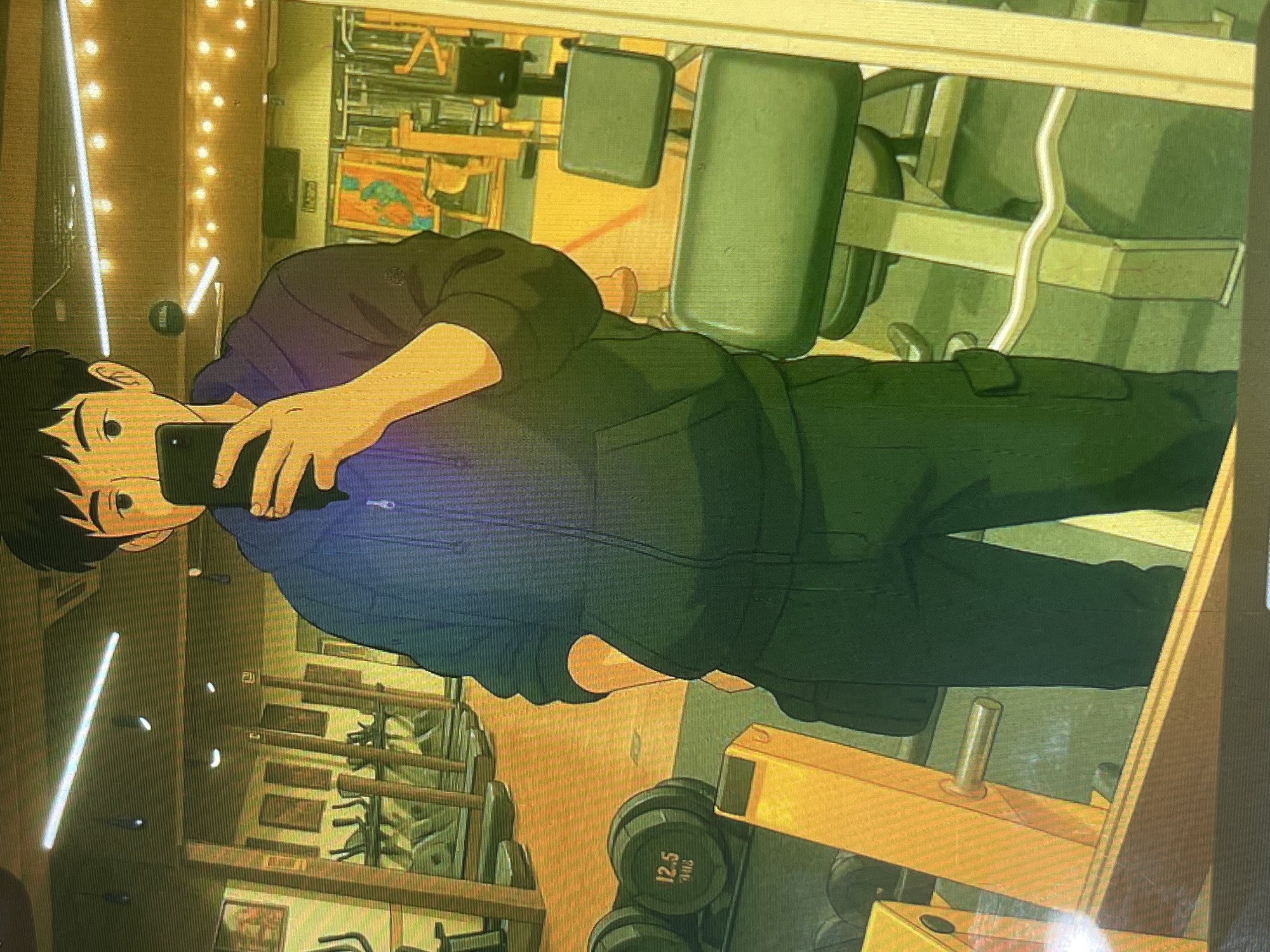 The image depicts a young man taking a selfie in a gym. He is wearing a dark hooded jacket and dark pants, with a black bag slung over his shoulder. In the foreground, there are dumbbells and gym equipment. In the background, treadmills are visible, along with mirrors reflecting the gym's interior. The lighting appears to be artificial, with overhead lights and string lights adding to the ambiance. The overall aesthetic is that of a typical fitness facility. The image does not contain any recognizable landmarks or elements that would indicate the location as Bishkek City, Kyrgyzstan.
