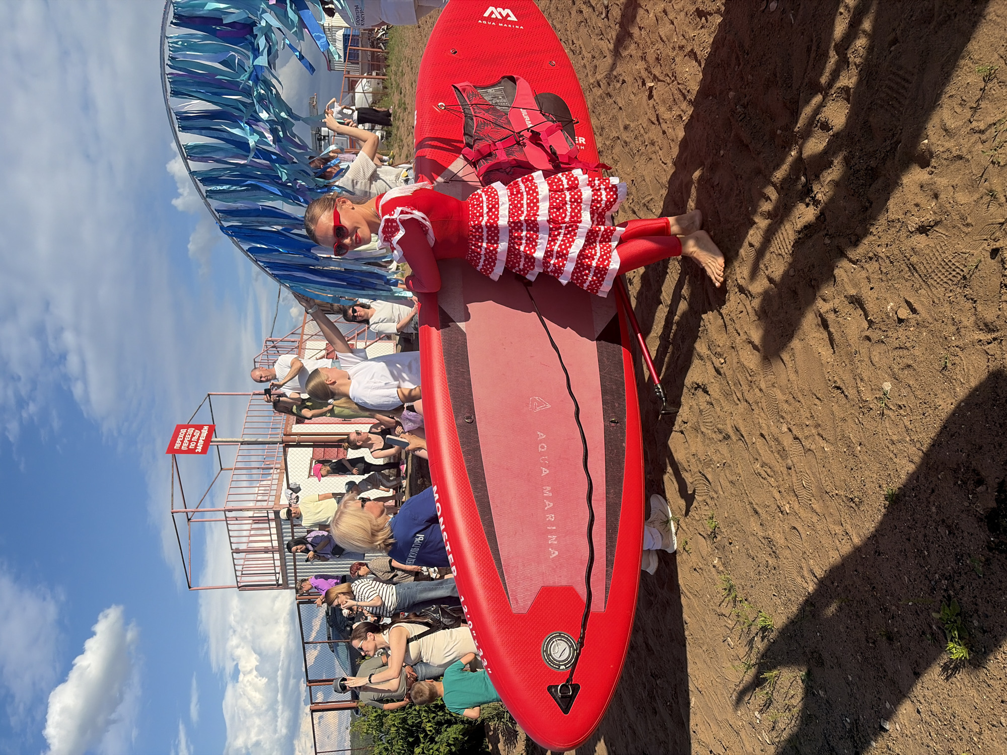 A young woman in a red polka-dot dress and red leggings stands on a sandy beach, holding a red paddleboard with "AQUA MARINA" written on it. She is wearing red sunglasses and smiling at the camera. Behind her, a crowd of people are gathered, some holding phones and cameras. In the background, there is a blue and white striped arch and a red sign with Cyrillic text. The sky is blue with white clouds. The scene appears to be taking place at the Chaikovsky City Beach in Chaikovsky, Russia, likely during a festival or event.