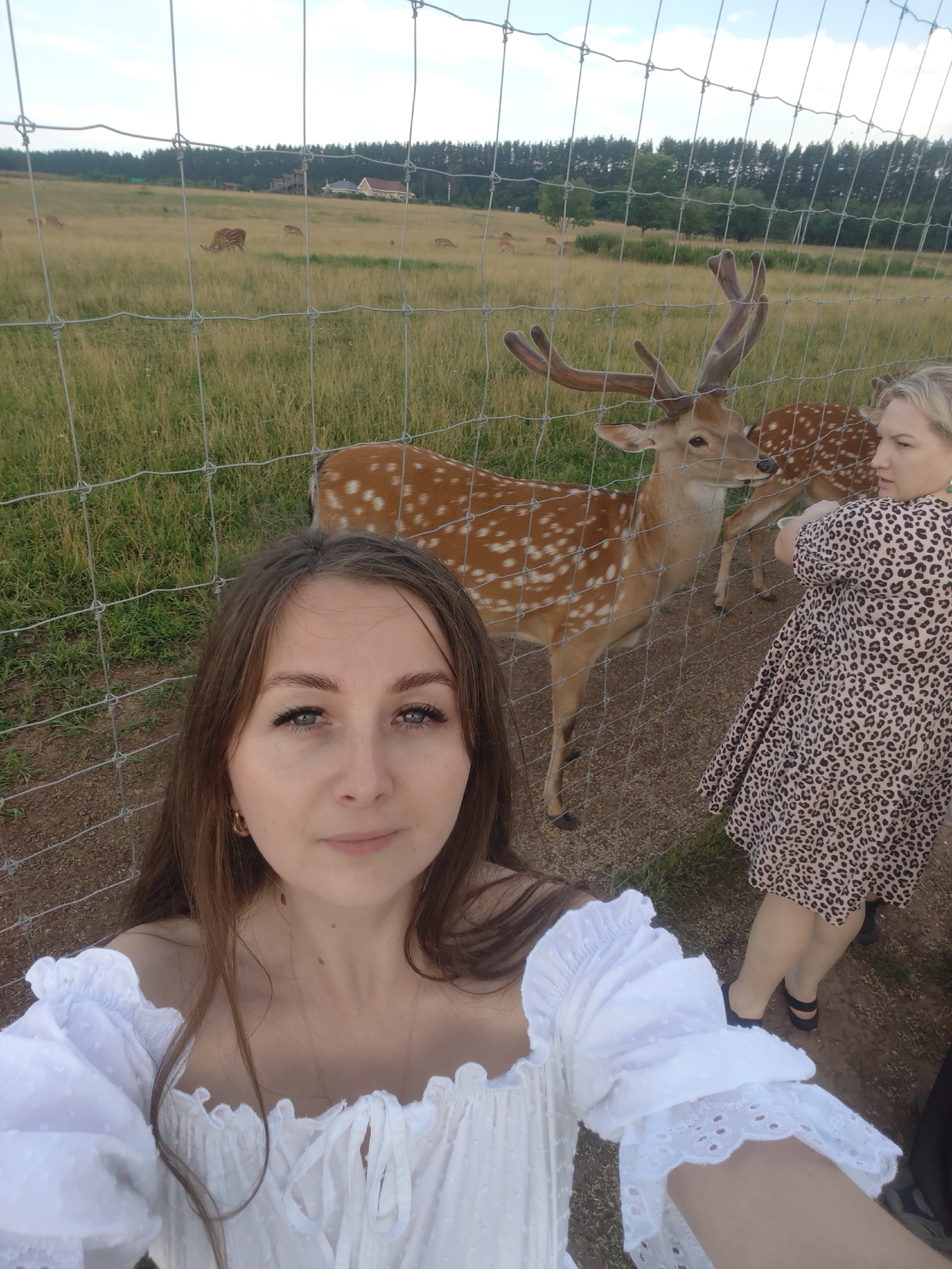 In a selfie taken at a zoo enclosure, two women are visible. The woman in the foreground, with long brown hair, is wearing a white off-the-shoulder top. To her right, another woman with blonde hair is wearing a leopard-print dress. Behind the women, a male deer with antlers is standing next to a fence. In the background, several other deer are grazing in a grassy field. A forest borders the field, and a few buildings are visible in the distance. The sky is cloudy.