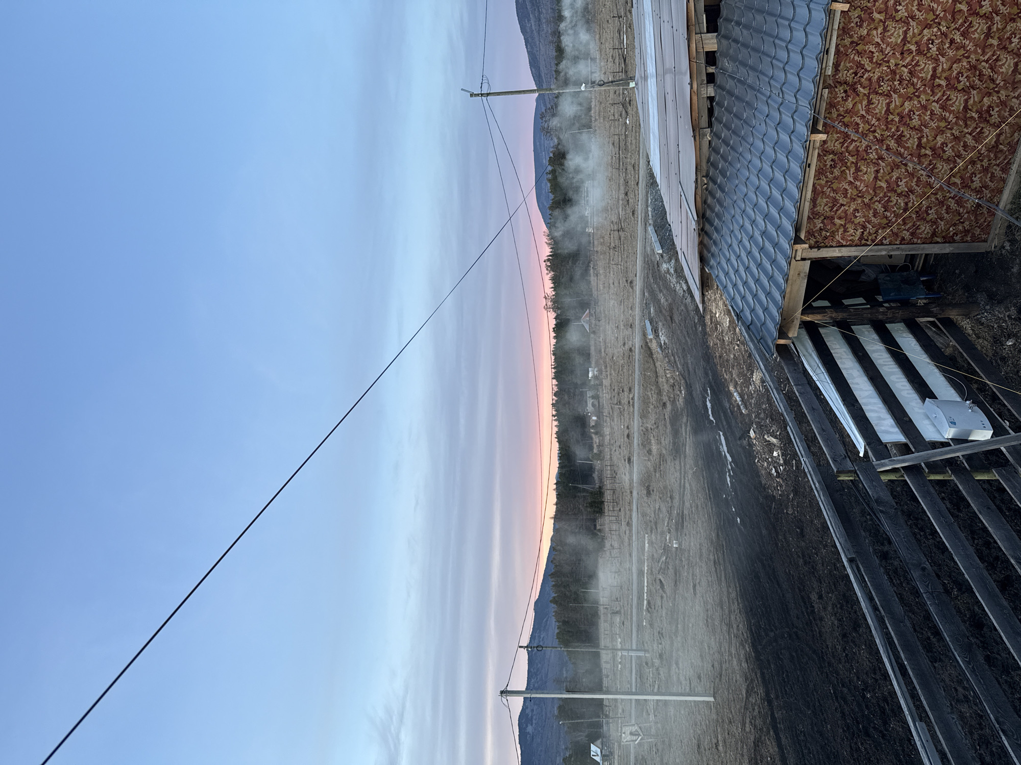 The image depicts a rural scene, possibly near the city of Tyulyuk, Russia. The sky is a gradient of blue and pink, suggesting dawn or dusk. Mountains are visible in the distance. In the foreground, there is an unpaved road or path, with a wooden structure to the right of the frame, partially covered with roofing materials. Utility poles and overhead wires are present, extending across the scene. Some mist or steam appears to be rising from the ground.