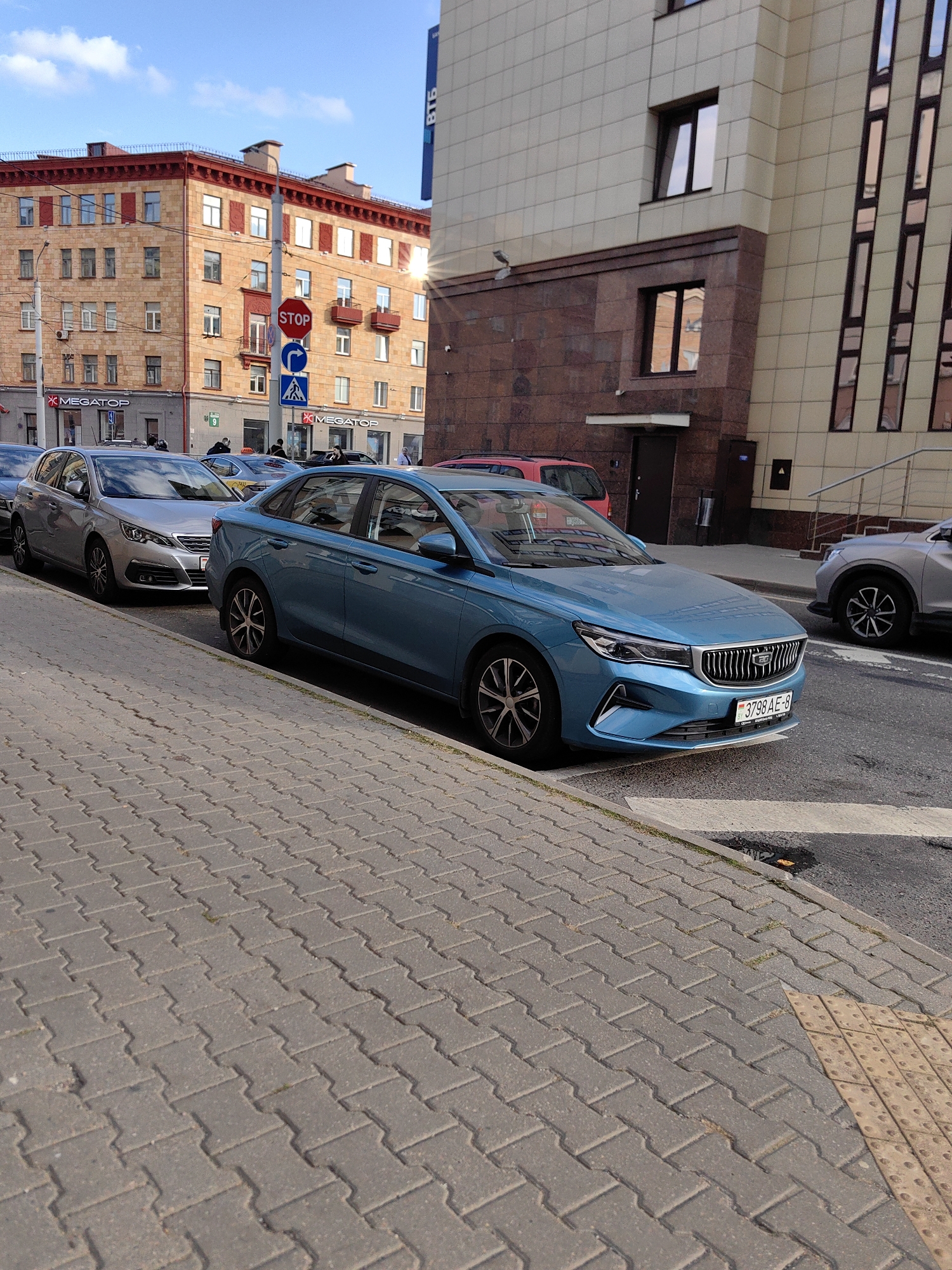 The image depicts a street scene in Minsk, Belarus, likely on a sunny day. In the foreground, a blue sedan is parked on the right side of the street, its front end angled towards the viewer. The car has a license plate that reads "3798 AE-8". To its left, a silver Peugeot sedan is also parked, and further back, another silver SUV is visible. The cars are parked along a sidewalk made of interlocking concrete pavers.
In the background, a modern office building with a beige and dark brown facade stands on the right. A red building with a red-tiled roof, featuring numerous windows and balconies, is visible on the left. Traffic signs are mounted on poles in front of the red building, including a red octagonal STOP sign, a blue circular sign with a white arrow, and a blue square sign with a white pedestrian symbol. The sky is blue with some clouds. The presence of a blue sedan with a Belarusian license plate, the architecture of the buildings, and the traffic signs all suggest the location is Minsk, Belarus.