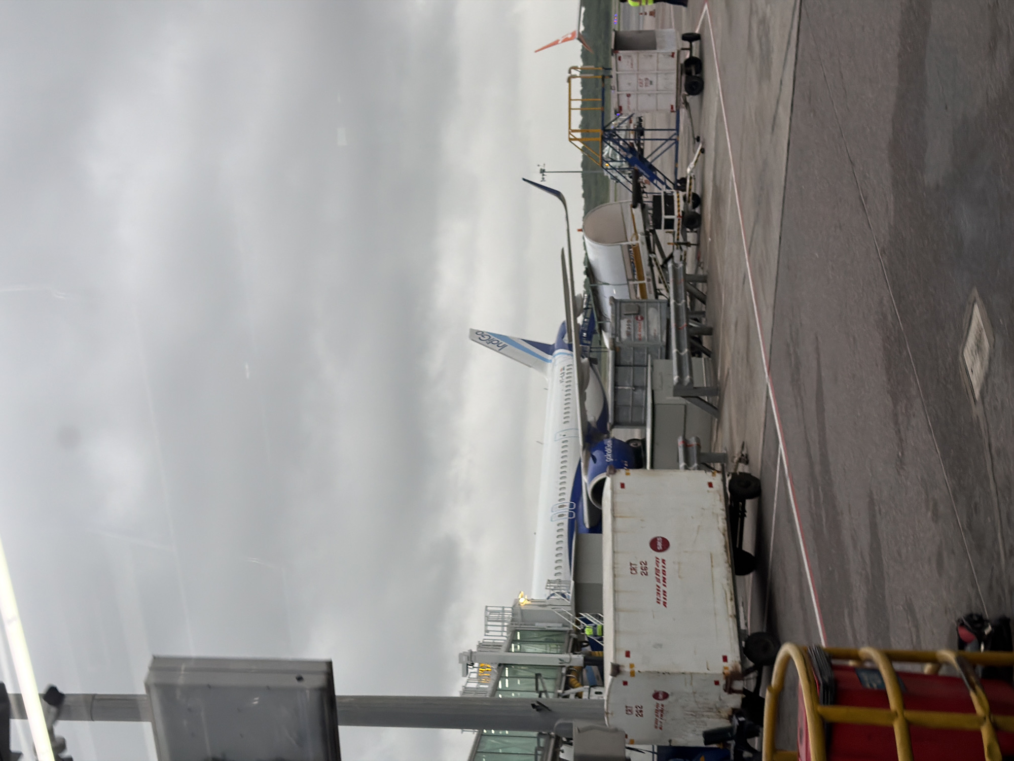 The image depicts an airplane parked at an airport gate, likely Rajiv Gandhi International Airport in Hyderabad, India, given the presence of Indigo and Air India branding. The sky is overcast, suggesting a cloudy day. In the foreground, a white cargo container with "AIR INDIA" and "CRT 262" written on it is visible. To the left, a yellow safety barrier encloses a red bin. Beyond the cargo container, a jet bridge connects to an Indigo Airlines airplane, identified by its blue and white livery and "IndiGo" branding on the tail fin. Another airplane, possibly also Air India due to the orange tail fin visible in the distance, is parked on the tarmac. Airport personnel in high-visibility vests are present near the aircraft and equipment. The scene is static, indicating the aircraft is preparing for or has just completed a flight.