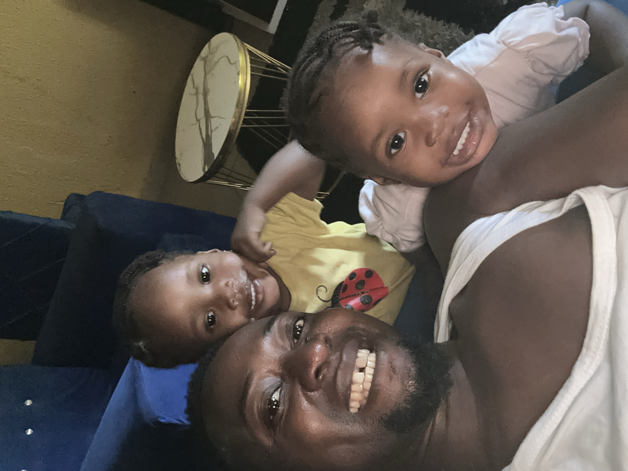 A selfie captures a man with two young children. The man is smiling broadly, revealing his teeth. One child is wearing a white shirt, and the other is wearing a yellow shirt with a ladybug on it. The background includes a gold-colored table and a blue sofa. The location appears to be indoors, possibly a home in the city of Unknown, Unknown.