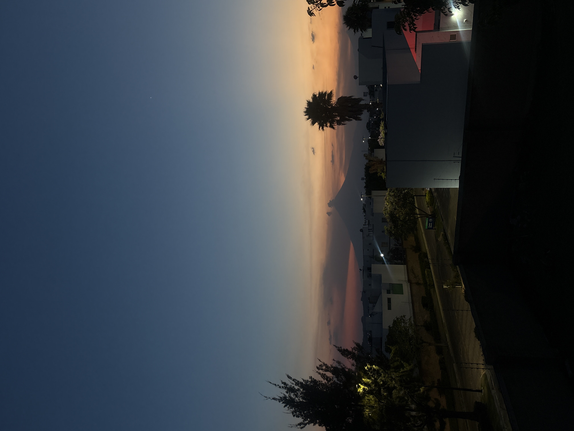 The image depicts a view in San Bernardino Tlaxcalancingo, Mexico, at dusk or twilight. The sky transitions from a deep blue in the lower-left to an orange-peach hue towards the upper-right. Buildings are visible on the right, with some illuminated by streetlights or building lights. Silhouetted trees, including a palm tree, are positioned between the sky and the buildings. A small, faint point of light, likely a star, is visible in the dark blue section of the sky. A street is present, illuminated by lampposts. The image has a vertical orientation.