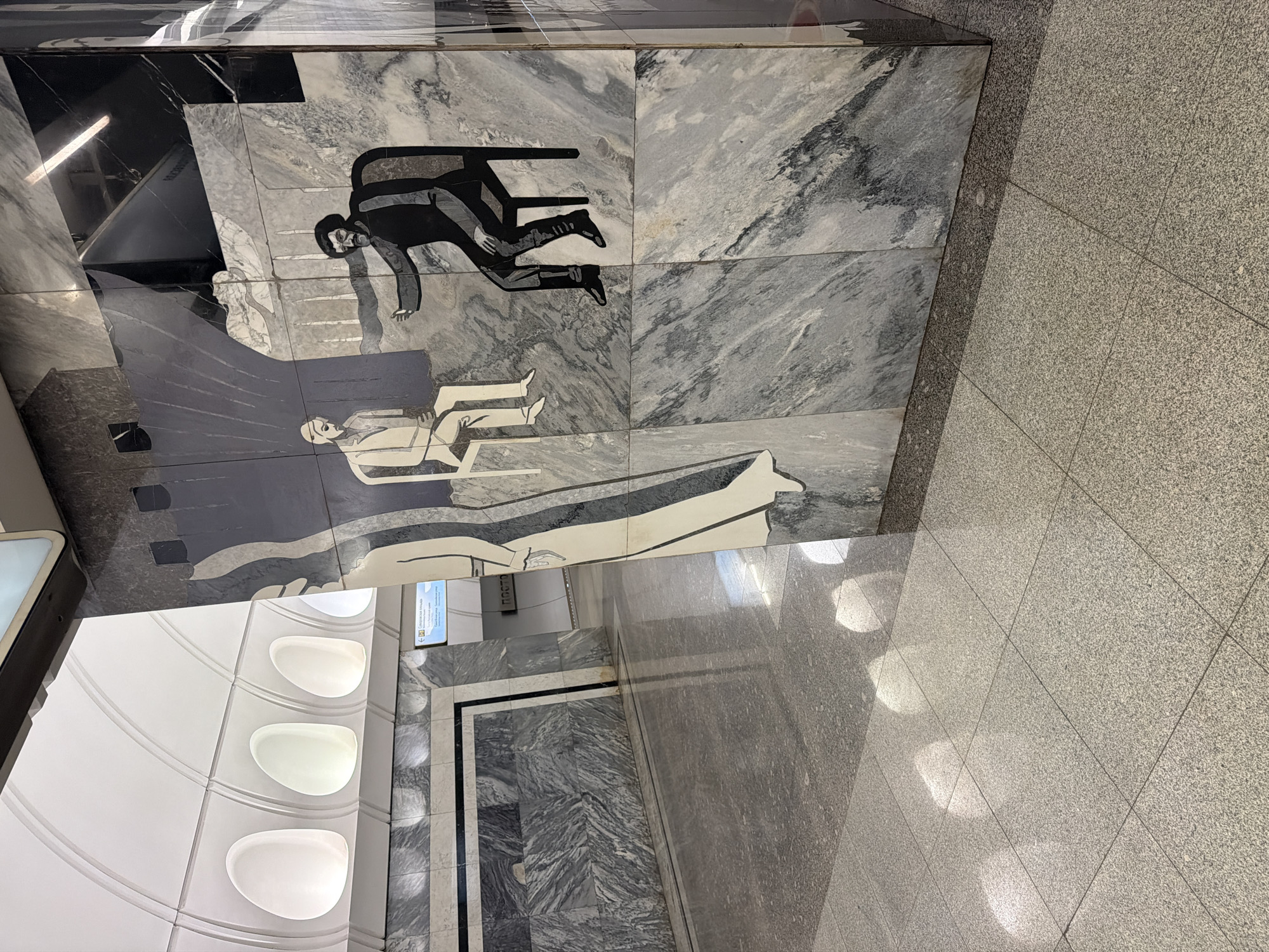 The image depicts the interior of a Moscow Metro station, specifically the Komsomolskaya station. The foreground is dominated by a polished granite floor reflecting light. To the right, a marble-clad pillar features a mosaic artwork depicting two figures. One figure, dressed in black, is seated on a chair and gesturing with an outstretched hand towards another figure seated opposite them, who is wearing a light-colored suit. The artwork is stylized and abstract, composed of geometric shapes and bold lines. In the background, a curved white ceiling with integrated lighting is visible, along with more marble paneling on the walls. A sign in Russian is partially visible, indicating directions within the station. The overall impression is one of clean lines, modern design, and artistic embellishment characteristic of Soviet-era metro architecture.