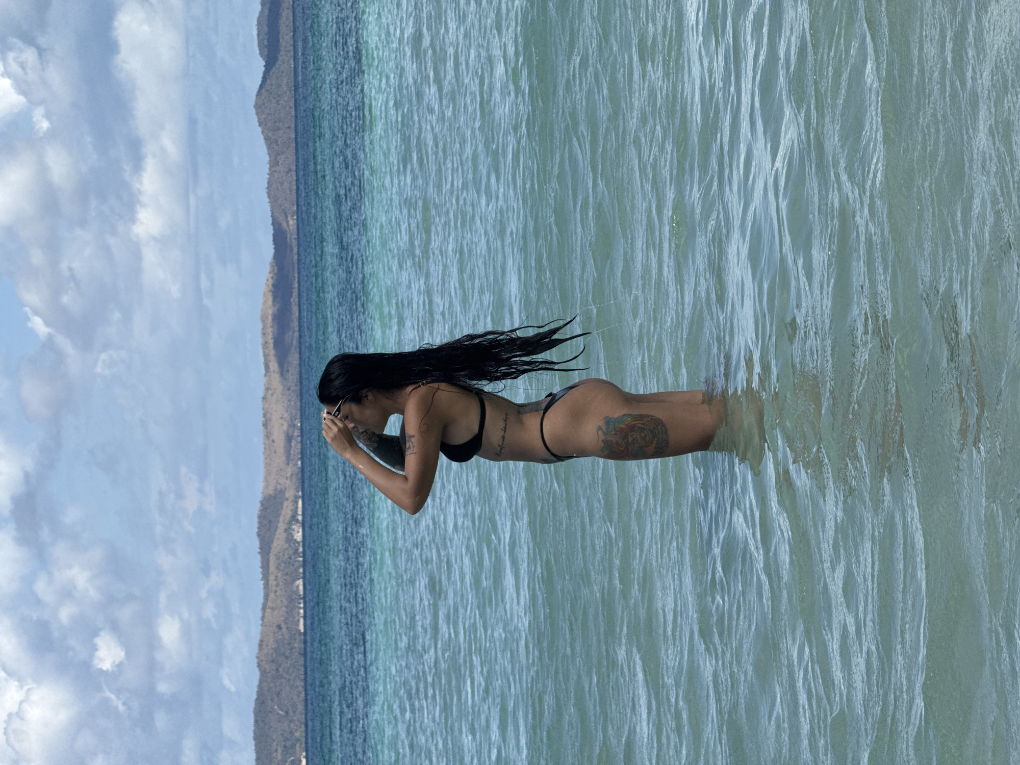 A woman stands in shallow water, wearing a black bikini, with her long dark hair wet. She is holding sunglasses near her face. The water appears clear and calm, and mountains can be seen in the background under a cloudy sky. The location is likely Pattaya City, Thailand, based on the prompt.