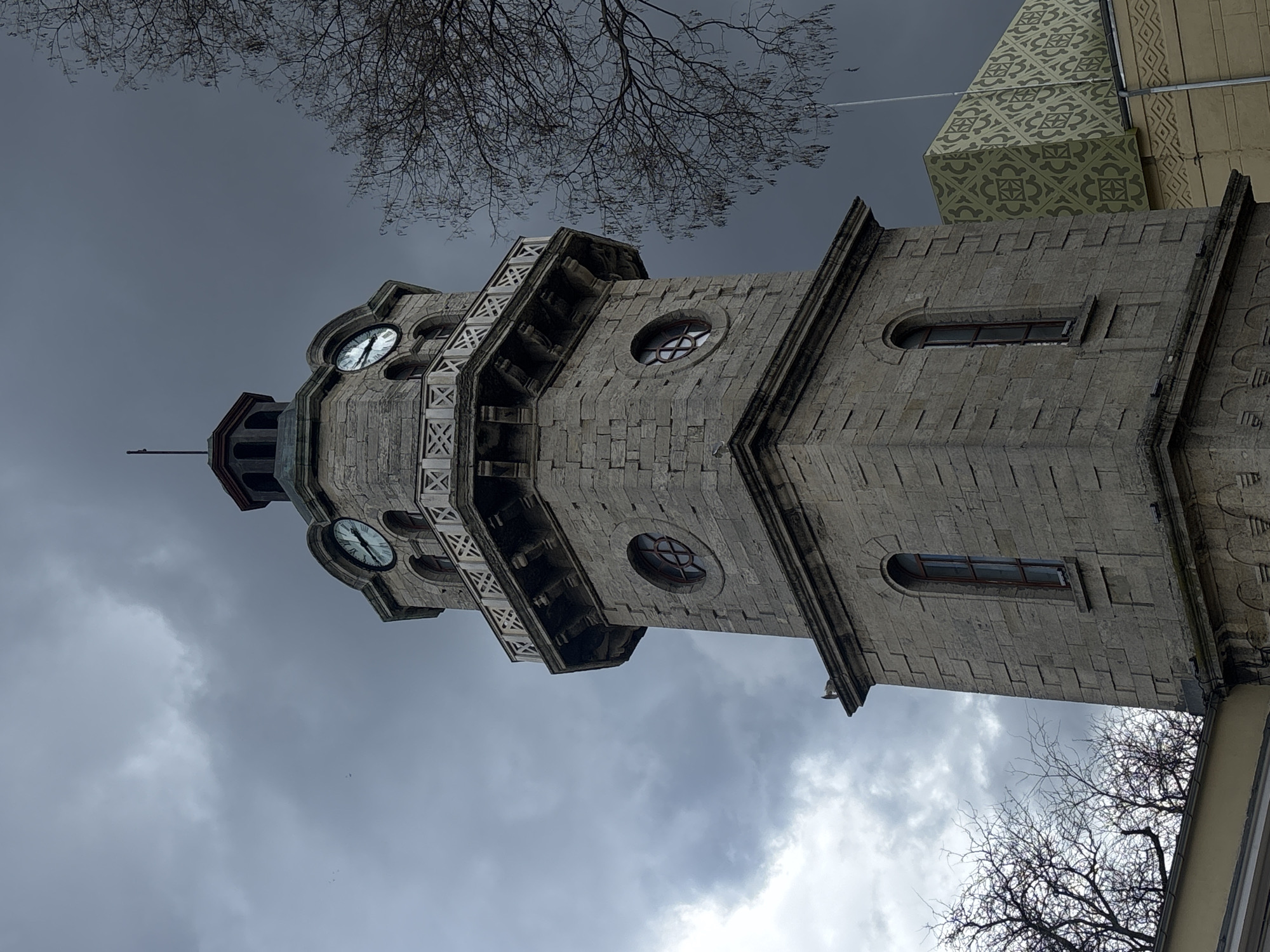 The image shows a low-angle view of the Varna Clock Tower in Varna, Bulgaria against an overcast sky. The stone tower features four clock faces, each visible on a different side. It has a multi-tiered structure with windows, including round and narrow rectangular openings. The tower has decorative detailing and a narrow spire atop. Bare tree branches are visible on either side of the tower against the cloudy sky. Part of an adjacent building with a patterned design is also visible.