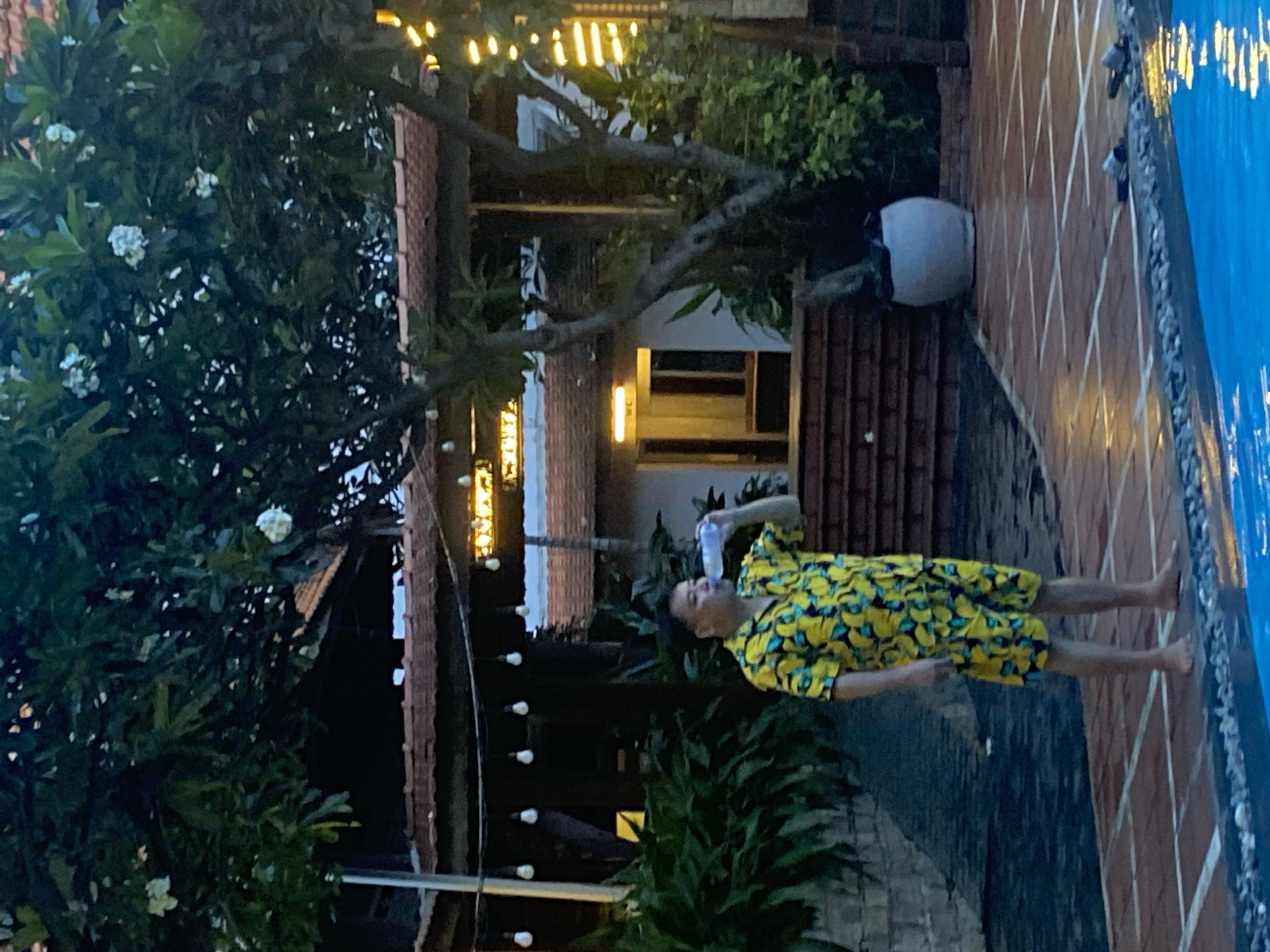 In Phu Thuy Ward, Vietnam, a man is standing by a swimming pool, drinking from a plastic bottle of water. He is wearing a bright yellow short-sleeved shirt and shorts adorned with a banana print. The background features a building with a tiled roof and several outdoor lights hanging from wires. Lush green foliage, including trees and bushes, surrounds the area. The ground around the pool is paved with red tiles.