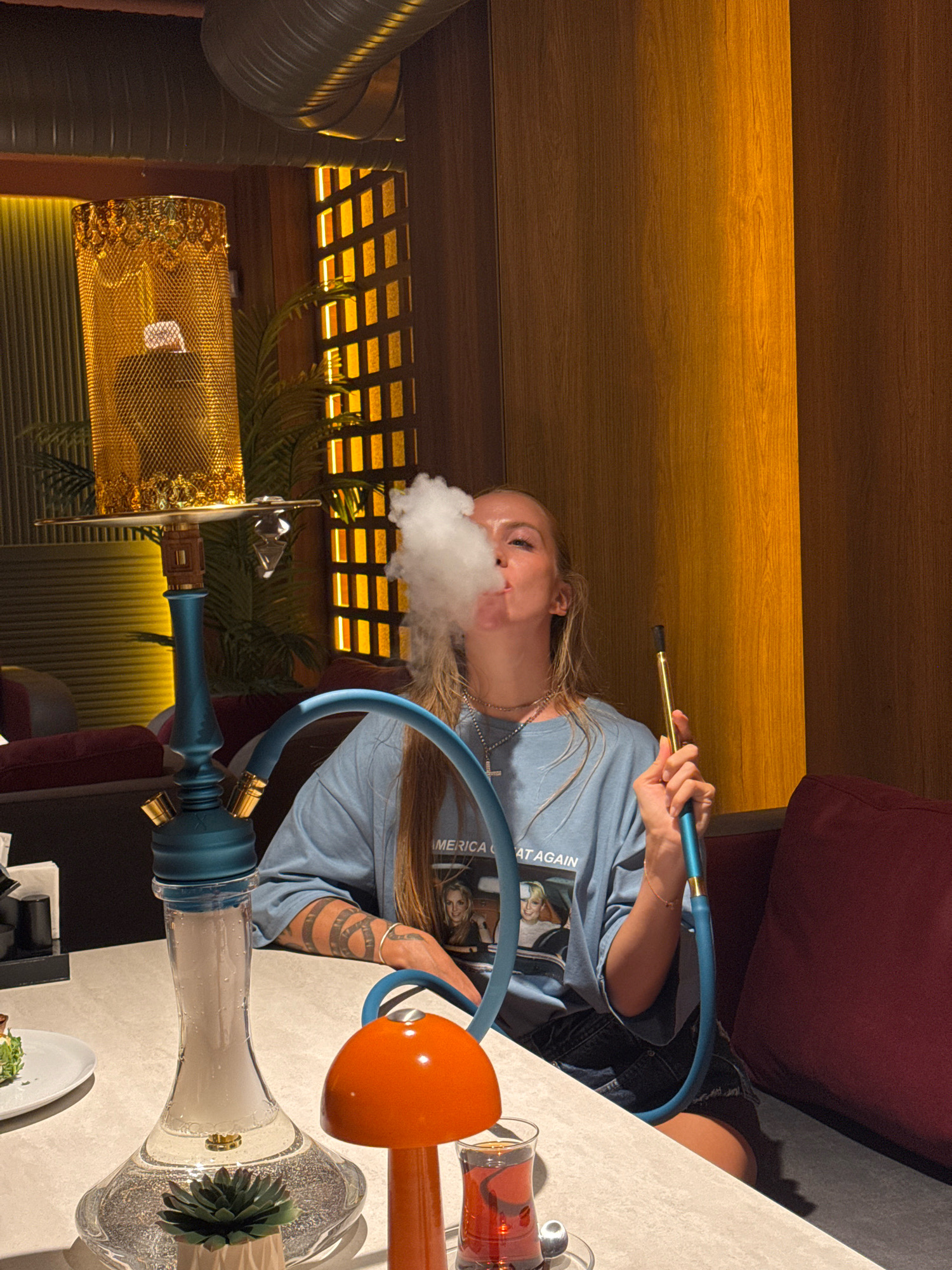 A young woman with blonde hair is exhaling smoke from a hookah pipe in what appears to be a modern lounge or restaurant in Antalya, Turkey. She is seated at a table, wearing a light blue t-shirt and denim shorts. The hookah, with its teal base and gold ornate top, is prominent on the table, alongside a small potted succulent and a glass of red tea. The background features warm wooden paneling and decorative lighting. The woman's arms are visible, showing intricate tattoos. The environment suggests a relaxed, contemporary setting.