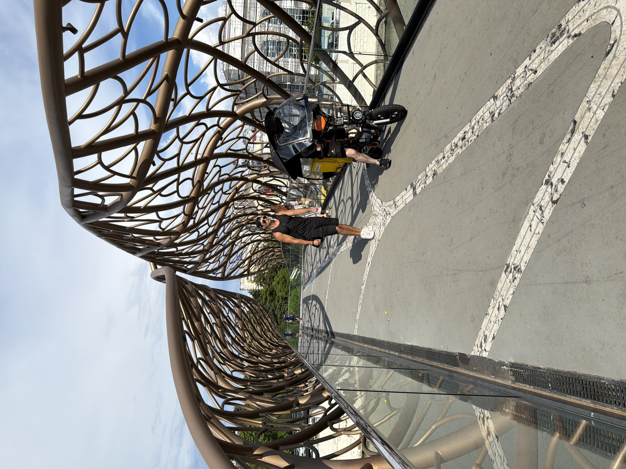 This is a wide, low-angle shot of a modern bridge with a unique, organic-looking metallic structure resembling intertwined branches or roots. The bridge has a walkway with a distinctive patterned surface, marked with white lines. A glass railing runs along one side, reflecting the metallic structure and the surroundings.
In the foreground, a man in a black tank top and shorts is standing on the walkway, facing the camera with a relaxed expression and wearing sunglasses. Behind him, a three-wheeled vehicle, possibly an electric scooter or a small enclosed motorcycle, is parked. The vehicle is black with a transparent canopy and has a yellow box attached to the back, suggesting it might be used for delivery.
Further down the walkway, two figures are visible in the distance, walking away from the camera. The setting appears to be outdoors, with a bright sky and some clouds visible above the bridge structure. Trees and buildings can be seen in the background, indicating an urban or park-like environment. The lighting suggests it is daytime, likely late morning or afternoon, with bright sunlight casting shadows. The overall mood is casual and leisurely, with the man in the foreground appearing comfortable and at ease. The location is identified as Sochi, Russia.