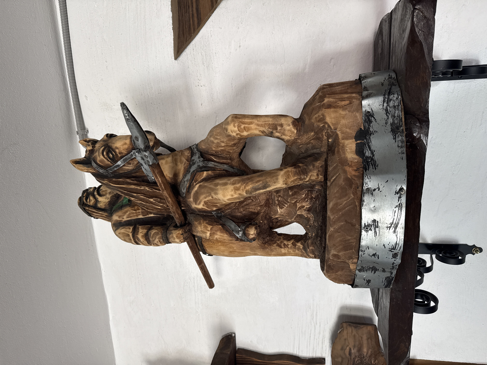 A close-up, eye-level shot shows a wooden sculpture of a man riding a horse, displayed on a dark wooden shelf against a white, textured wall. The sculpture is intricately carved, depicting the rider with a beard and wearing traditional clothing, holding a spear. The horse is also detailed, with a bridle and saddle visible. The base of the sculpture is adorned with a metallic band. The shelf is supported by black, ornate metal brackets. To the left and right of the main sculpture, other wooden carvings are partially visible, suggesting a collection or display. The lighting is soft, highlighting the textures of the wood and the wall. The scene appears to be indoors, possibly in a home or gallery.