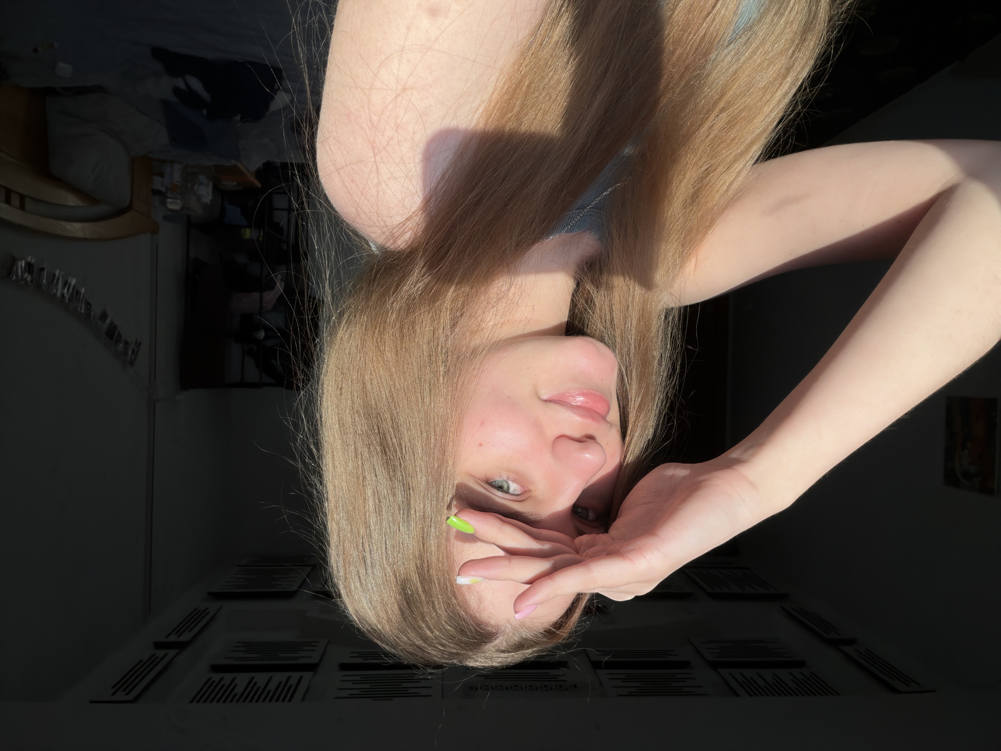 A young woman with long blonde hair is looking into the camera, shielding her eyes with her right hand. She is wearing a grey top. The lighting suggests it is late afternoon, with sunlight casting long shadows. The background is dark and appears to be an indoor setting, possibly a bedroom, with a bed visible on the right and some items on shelves on the left. There are acoustic panels on the ceiling. The location is not specified as Dnipro, Ukraine.