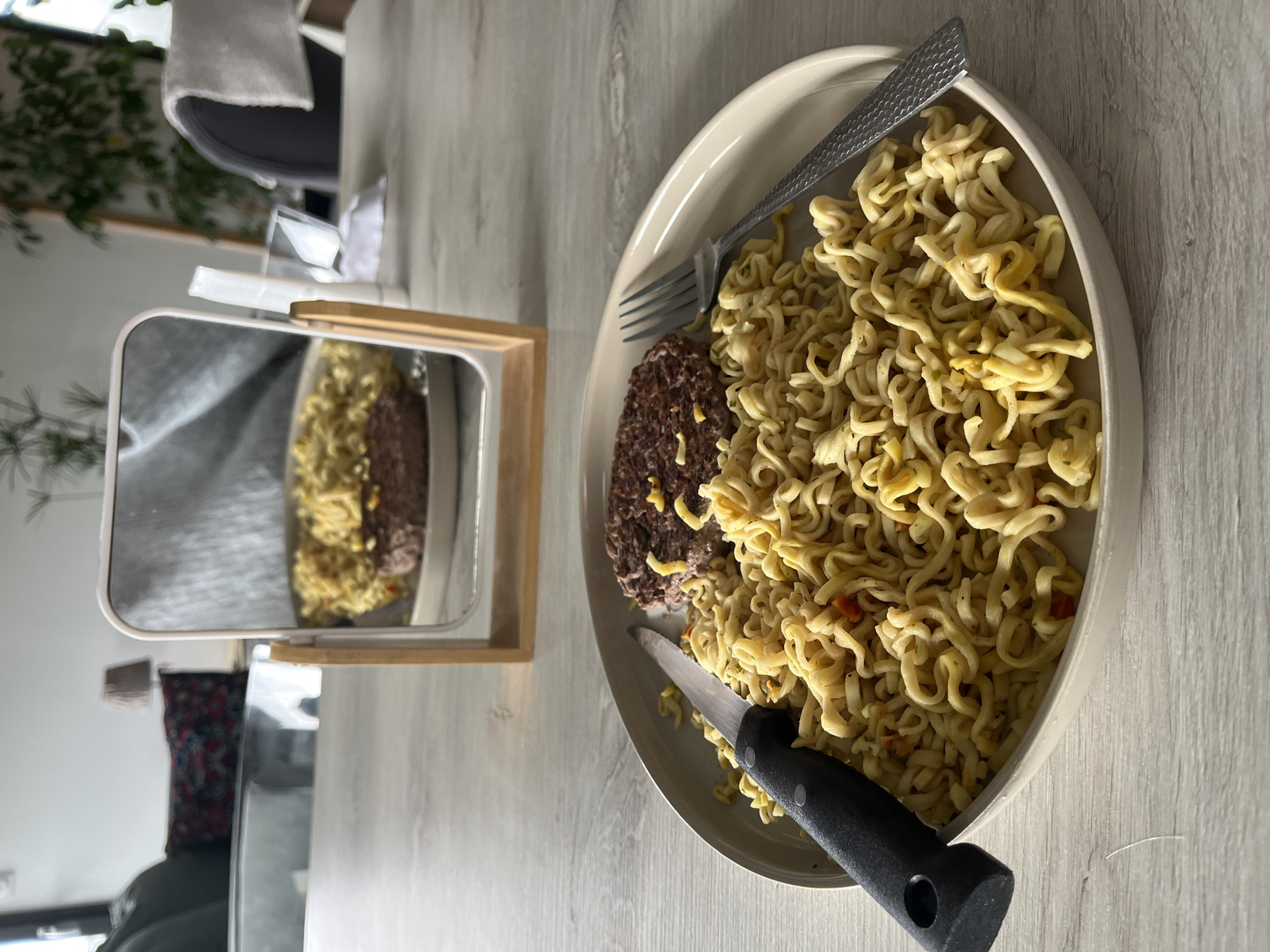 This image, captured in Chambœuf, France, depicts a meal set on a light gray wooden-grain table within an indoor residential environment. A round, off-white ceramic plate occupies the central foreground, containing a cooked hamburger patty and a quantity of instant ramen noodles, seasoned with flecks of green and orange. A black-handled knife with a serrated blade rests on the left side of the plate, partially atop the noodles. A silver-colored fork with a textured handle is positioned on the right side, also partially over the noodles. Directly behind the plate, a small, white-framed mirror with a light wooden base stands upright, reflecting a distorted, inverted view of the meal, specifically showing the noodles prominently on the burger. The background is out of focus, revealing elements of a living area: to the left, a dark sofa with a patterned cushion and a table lamp with a dark, textured shade are visible; to the right, green foliage from a houseplant and a dark chair with a white cloth draped over its back are discernible against white walls. No individuals are present in the scene.