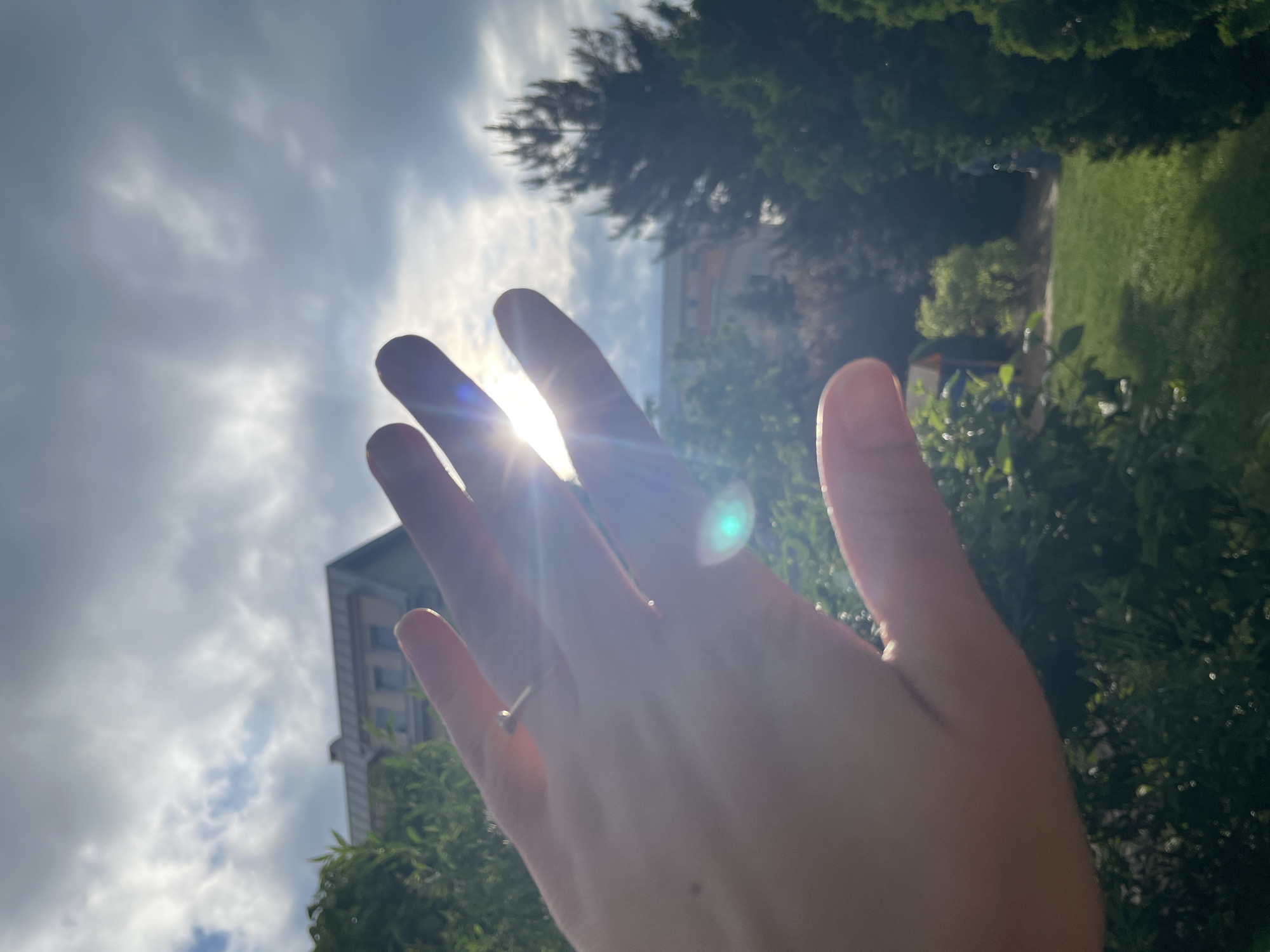An outdoor, upward-angled shot captures a human hand in the foreground, raised towards a bright sun. The sun's rays stream through the fingers, creating starburst effects and distinct lens flares, one of which is a prominent greenish-blue orb. A simple ring, possibly silver or white gold, with a small, round stone, is worn on the ring finger.
The background reveals a mix of natural and architectural elements. To the right, lush green foliage, including trees and bushes, fills the frame, suggesting a vibrant garden or park. Further back on the right, a patch of green grass is visible. On the left side, partially obscured by the hand and trees, stands a multi-story building with visible windows, indicating an urban or suburban setting. The sky above is partly cloudy, with bright patches where the sun shines through and areas of soft gray clouds. The strong sunlight suggests it is daytime, likely mid-morning or afternoon. No text is visible in the image.