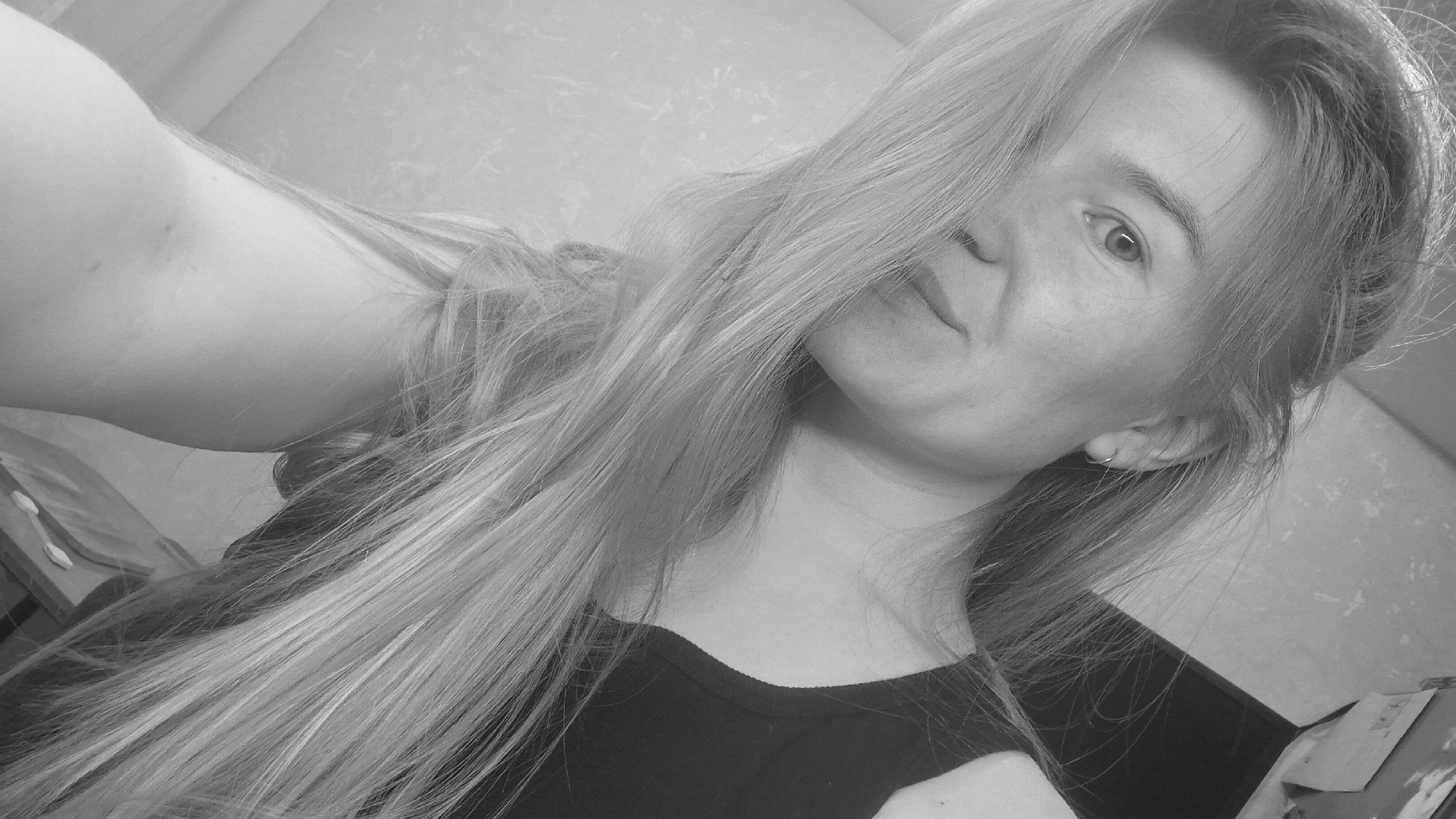 A grayscale, tilted selfie features a young woman with long, light-colored hair partially obscuring her face. Her right eye is visible, and she has freckles on her face. She is wearing a dark-colored top. In the background, a wall with a patterned texture can be seen. Her right arm is extended towards the camera. Some objects are visible on a surface to the left. The location is unknown.