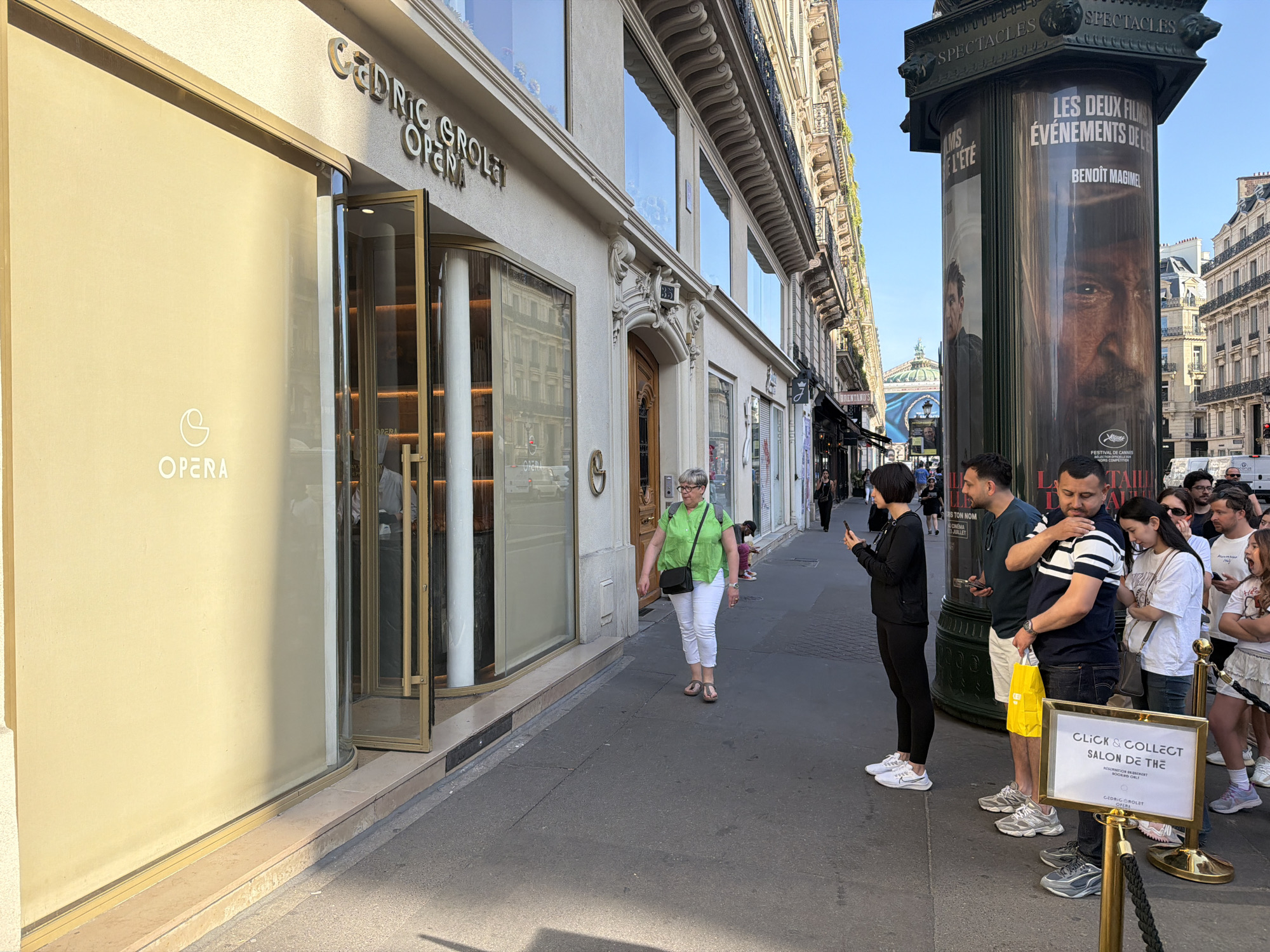 The image captures a street scene in Paris, France, featuring a queue of people waiting outside a shop with the name "CEDRIC GROLET OPERA" above the entrance. A large advertisement pillar stands beside the queue, displaying a movie poster of Benoit Magimel. A woman in a green shirt and white pants is walking on the sidewalk towards the shop. In the background, buildings line the street. The light indicates it's daytime. A sign reads "CLICK & COLLECT SALON DE THE".