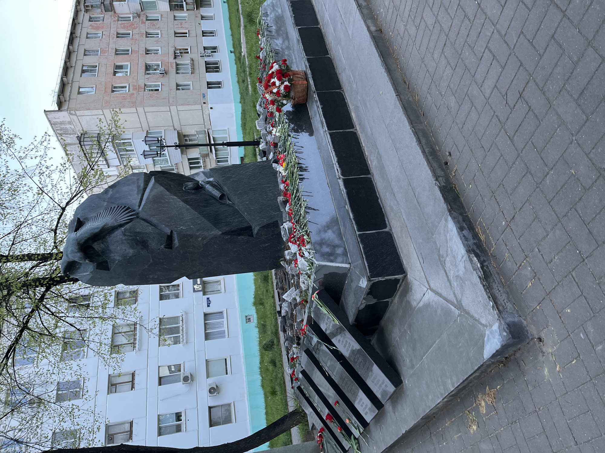 The image depicts a monument located in Chaikovsky, Russia. The monument features a large, dark-colored sculpture of a human head, possibly a memorial or commemorative figure. Flowers, including a basket of red and white flowers and individual red carnations, have been laid at the base of the monument. Behind the monument are several apartment buildings. The surrounding area is paved with gray brick. The sky is overcast, suggesting a cloudy day.