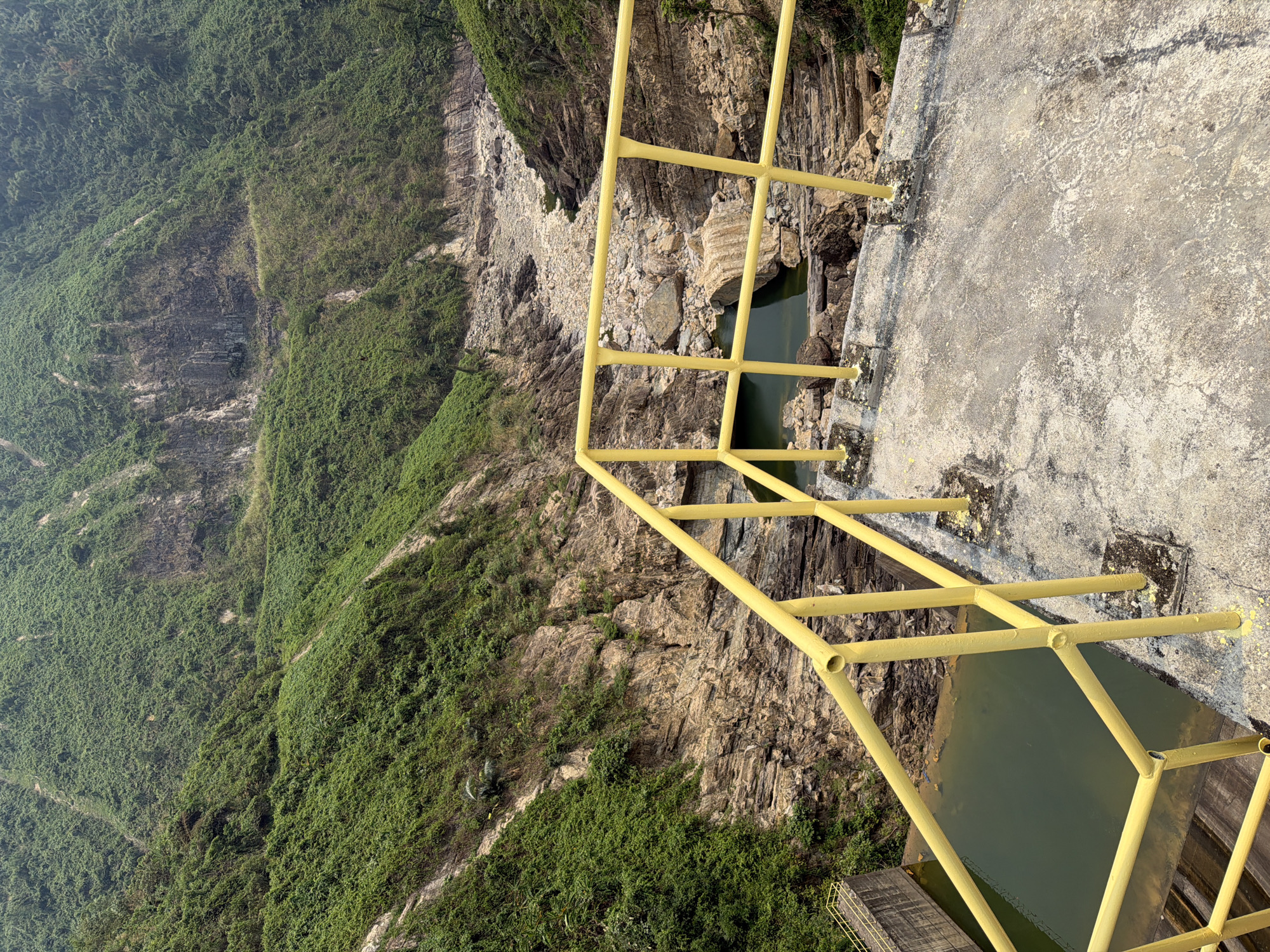 The image presents an elevated view from a concrete structure, likely a dam or overlook, featuring a bright yellow safety railing. Below, a steep, rugged cliff face descends into a body of water, possibly a reservoir. The cliff is partially covered with dense green vegetation, interspersed with exposed rock formations. The concrete platform shows signs of weathering. The location is likely in a mountainous or hilly area within the city of Đà Nẵng, Vietnam, considering the landscape and vegetation. No people are visible in the image. The focus is on the landscape and infrastructure.