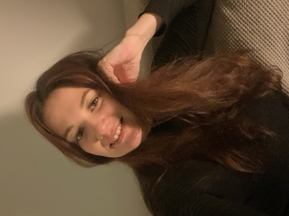 A woman reclines, her head tilted to the side and face angled upwards, smiling at the camera. Her hand is near her head, possibly touching her hair. She has auburn-brown hair and fair skin. She is wearing a dark-colored top. The background includes a pale-colored wall on one side and a textured sofa or chair on the other. Lighting seems soft and muted.