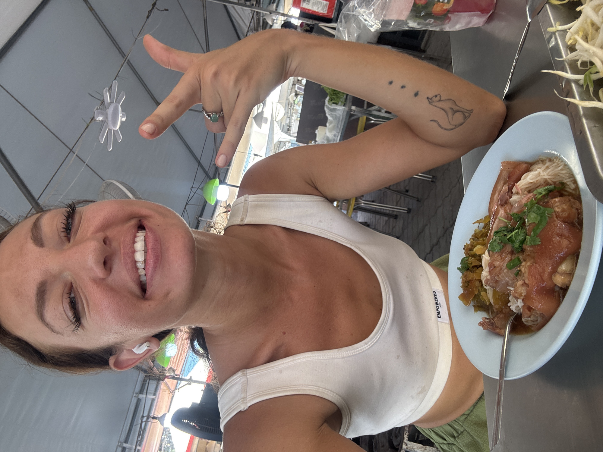 A woman is captured in a selfie, smiling broadly at the camera while making a hand gesture. She is wearing a white sports bra with the word "EMPOWERED" visible on the band on her left side, and green pants. A white earbud is in her right ear. Her right arm is raised, with the thumb and pinky extended in a "shaka" or "hang loose" gesture. On her right arm, she has a tattoo of a stylized cat-like figure in motion, with three small dots above it. A ring with a green stone is on her right ring finger.
She is seated at a metal table, with a light blue plate of food in front of her. The dish appears to be a Thai meal, possibly Khao Kha Mu (stewed pork leg), featuring tender pork, white rice, green herbs (likely cilantro), and pickled vegetables. A metal fork is resting in the food. To the right of the plate, a container holds a pile of fresh bean sprouts.
The setting appears to be an outdoor or semi-outdoor food stall or market in บ้านบางรักษ์, Thailand. Overhead, a light grey tent structure with metal beams and wires is visible, along with an overhead fan and a decorative, star-shaped light fixture. The lighting suggests it is daytime, and the woman's expression conveys happiness.