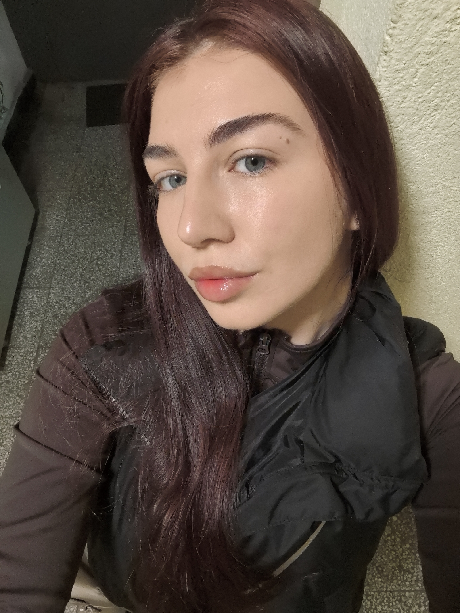 A young woman is photographed at close range in an indoor setting, seemingly near an exterior wall. The floor appears to be tiled. The woman has fair skin, light-colored eyes, and dark reddish-brown hair. She is wearing a dark brown jacket or long-sleeved shirt, and a black vest. The location is potentially Stara Zagora, Bulgaria, but this cannot be confirmed from the image. No specific actions or interactions are discernible.