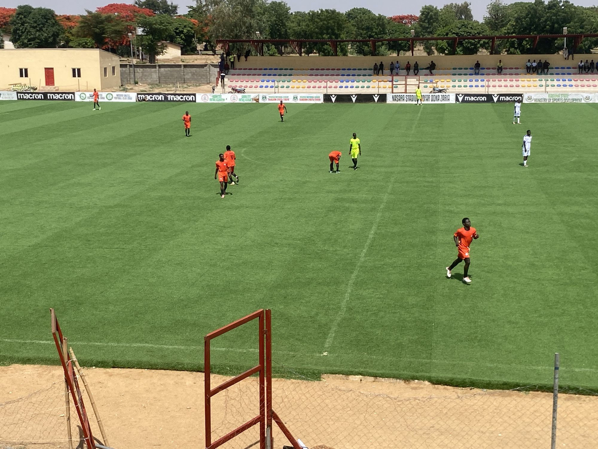 The image shows a soccer field in Tudun Wada, Nigeria. Players in orange and white uniforms are scattered on the green field. One player wears a green and yellow uniform. Banners with logos and text that include "macron", "NASARA STADIU", "KADUNA STATE" and "MINISTRY OF SPORTS DEVELOPMENT" are visible along the sidelines. Spectators are seated in the stands in the background, sheltered by a roofed structure. A building with a red door and wall borders the field. Trees and a light blue sky are visible above the buildings. A red goal frame and a sandy area with a wire fence are in the foreground.