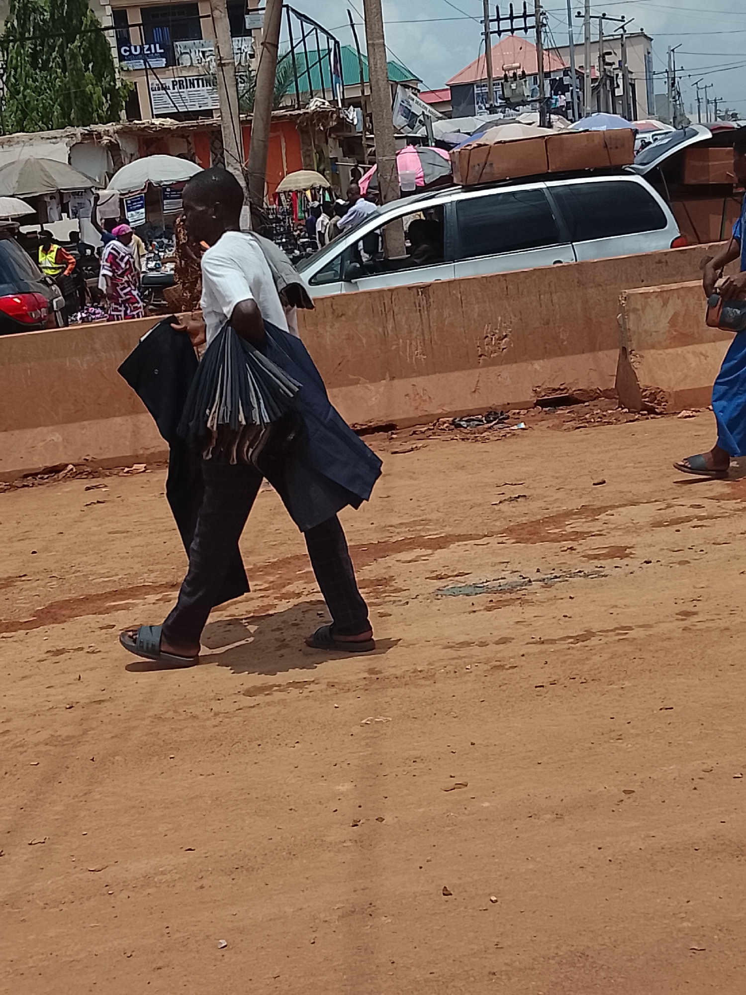 The image shows a man walking on a dusty, brown-colored ground surface in an urban area. He is wearing a white shirt, dark-colored pants, and sandals, and he is carrying a dark-colored garment or object in his left hand and what appears to be a bundle of umbrellas or similar items in his right. Behind him are indications of a bustling street scene, including people, vehicles, and vendor stalls with umbrellas. A light-colored minivan with boxes or items stacked on its roof is partially visible, as is a section of a concrete barrier. The background includes buildings, utility poles, and various signs. The location is identified as Katambe, Nigeria.